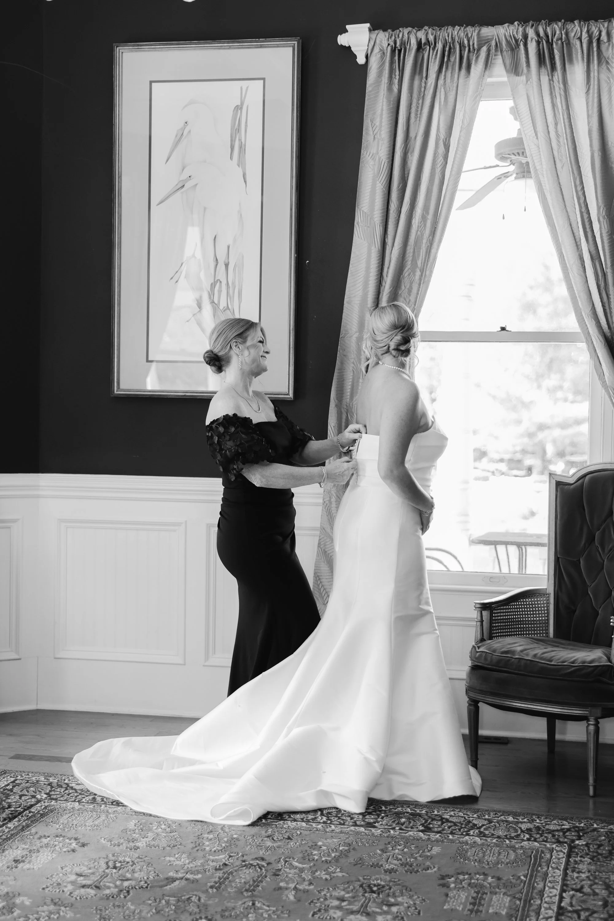 A woman in a strapless wedding gown standing by a window as another woman, possibly her mother, adjusts her belt or sash. The scene takes place in a well-decorated room with a large framed painting of herons on the wall and curtains framing the windo
