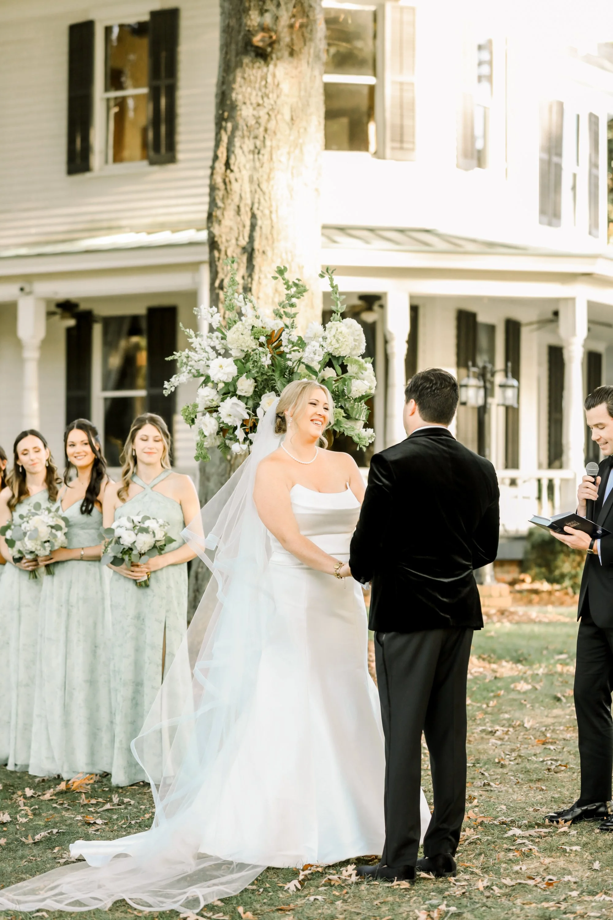 A wedding ceremony outdoors with the bride and groom holding hands, surrounded by bridesmaids holding bouquets, with a large floral arrangement and a house in the background, during daylight.