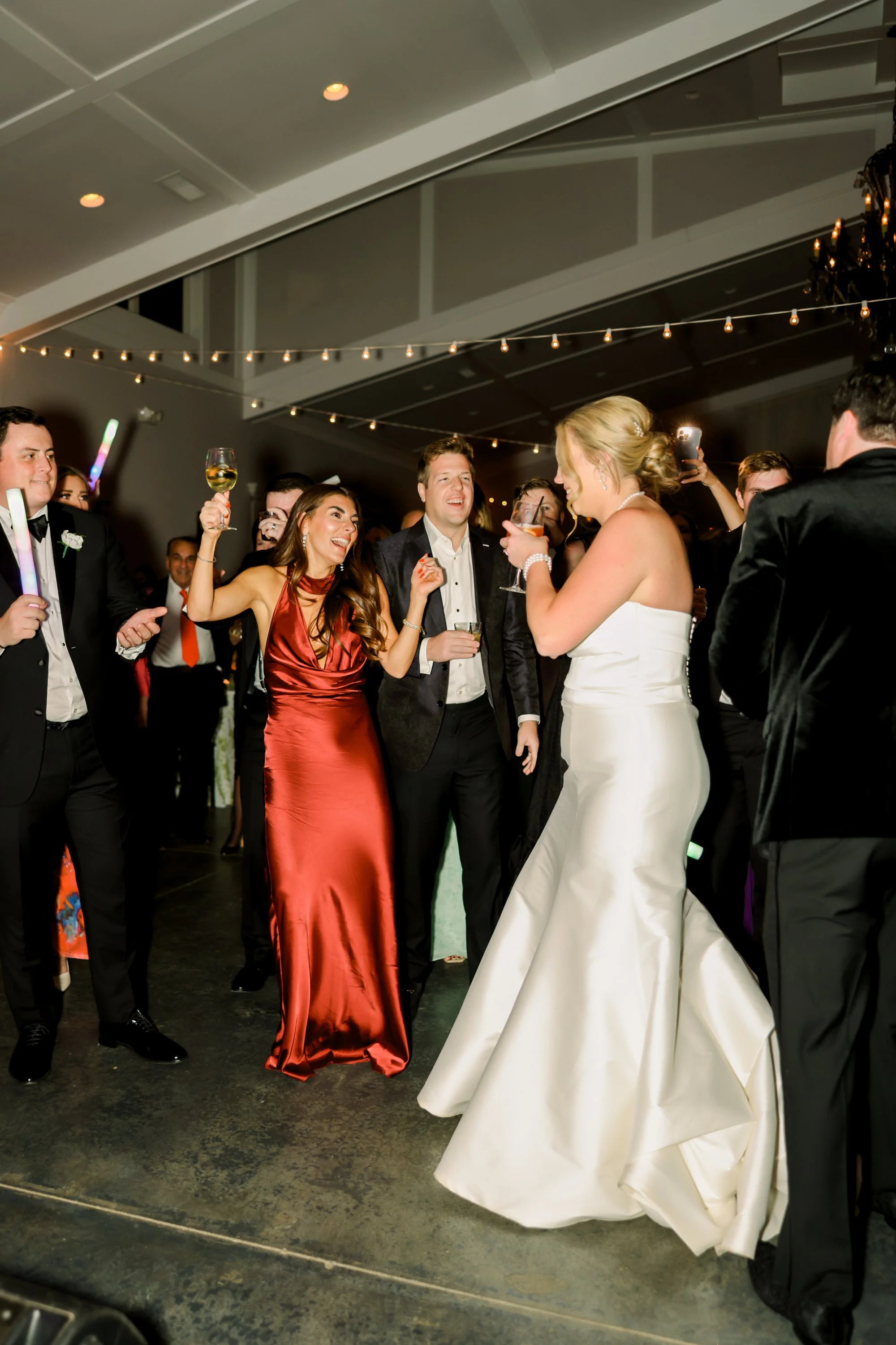 Group of people dancing and celebrating at a wedding reception, including a woman in a white wedding gown and others in formal attire, with drinks in hand.