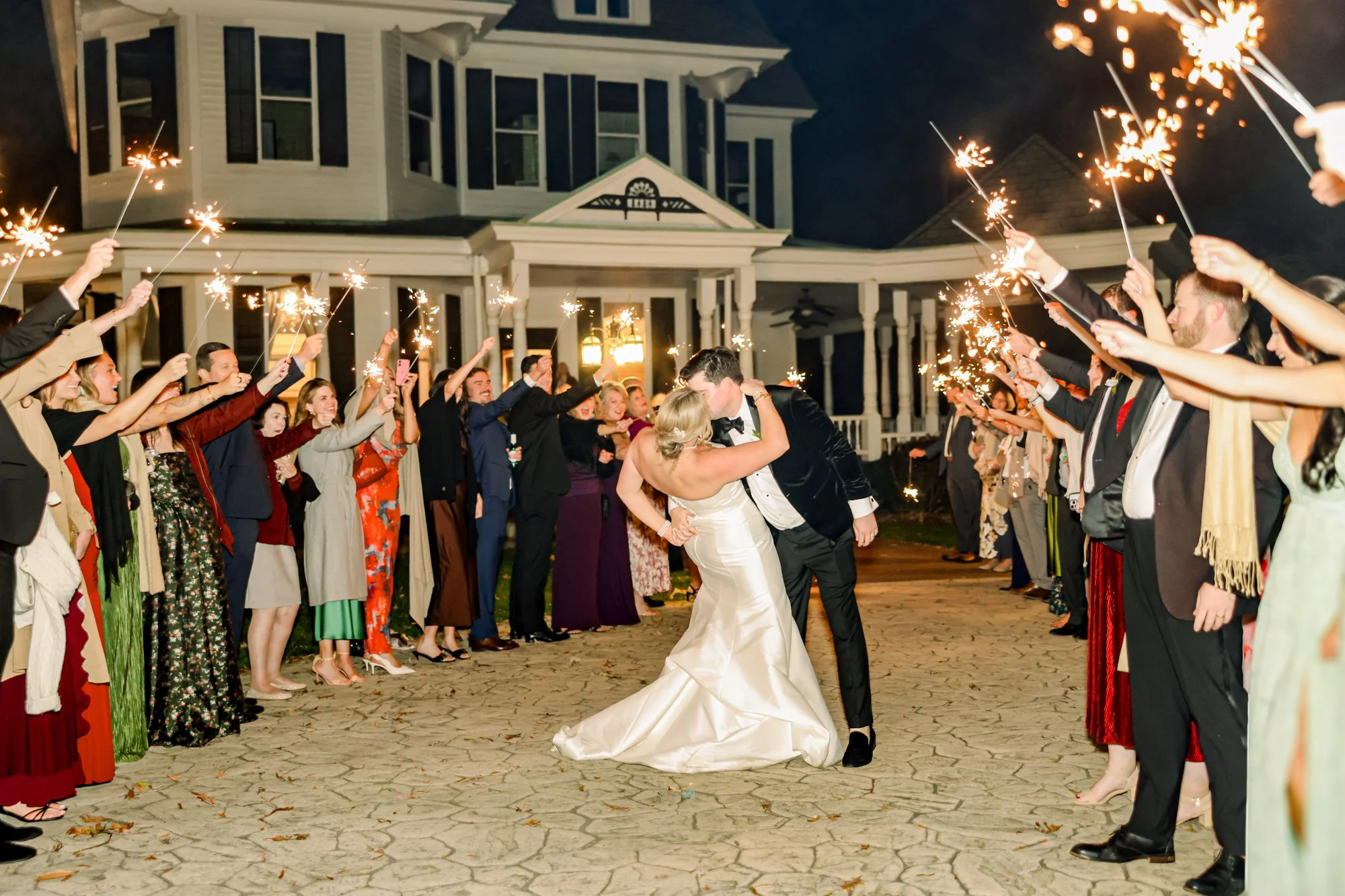 Bride and groom kissing while surrounded by guests holding sparklers outside a house at night.