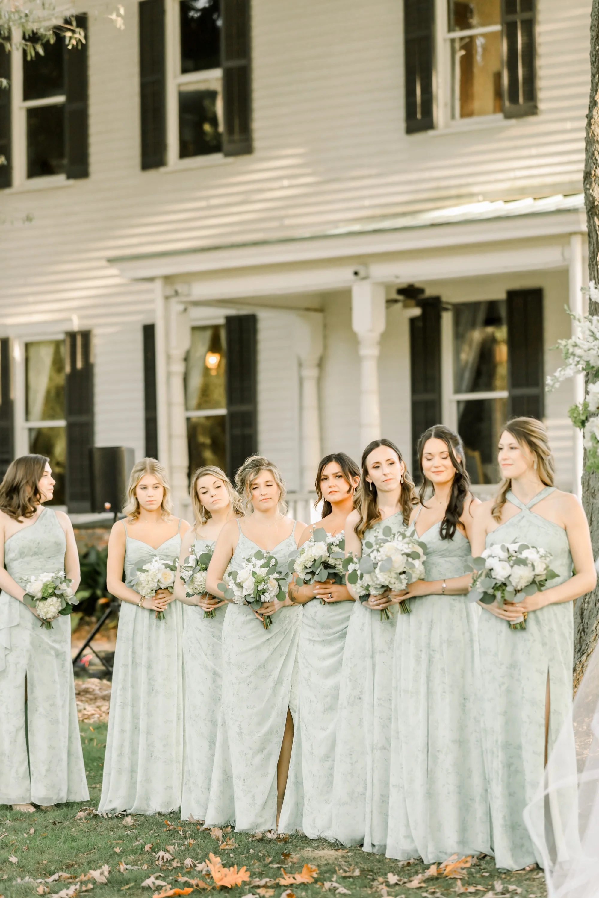Group of eight women in matching green dresses holding bouquets, standing outdoors in front of a white house with black shutters, during what appears to be a wedding or formal event.