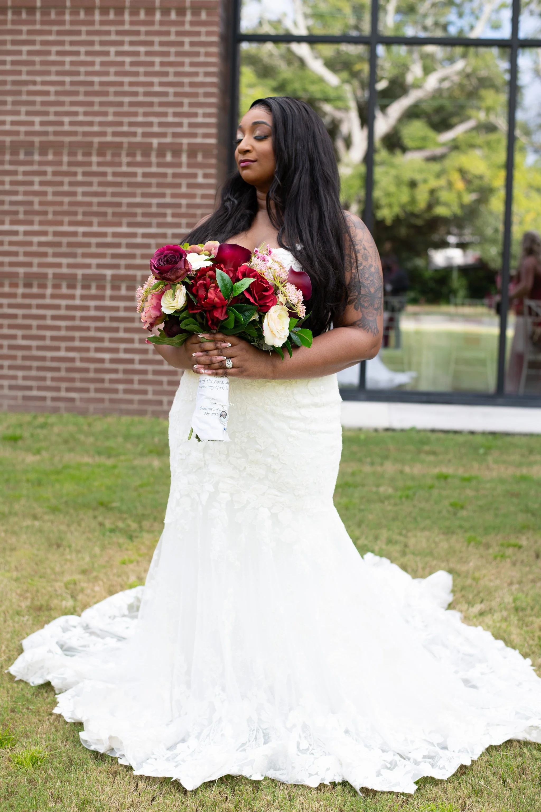 A bride in a white lace wedding dress holding a bouquet of red, white, and pink flowers, standing outdoors in front of a brick building and a window reflecting trees.