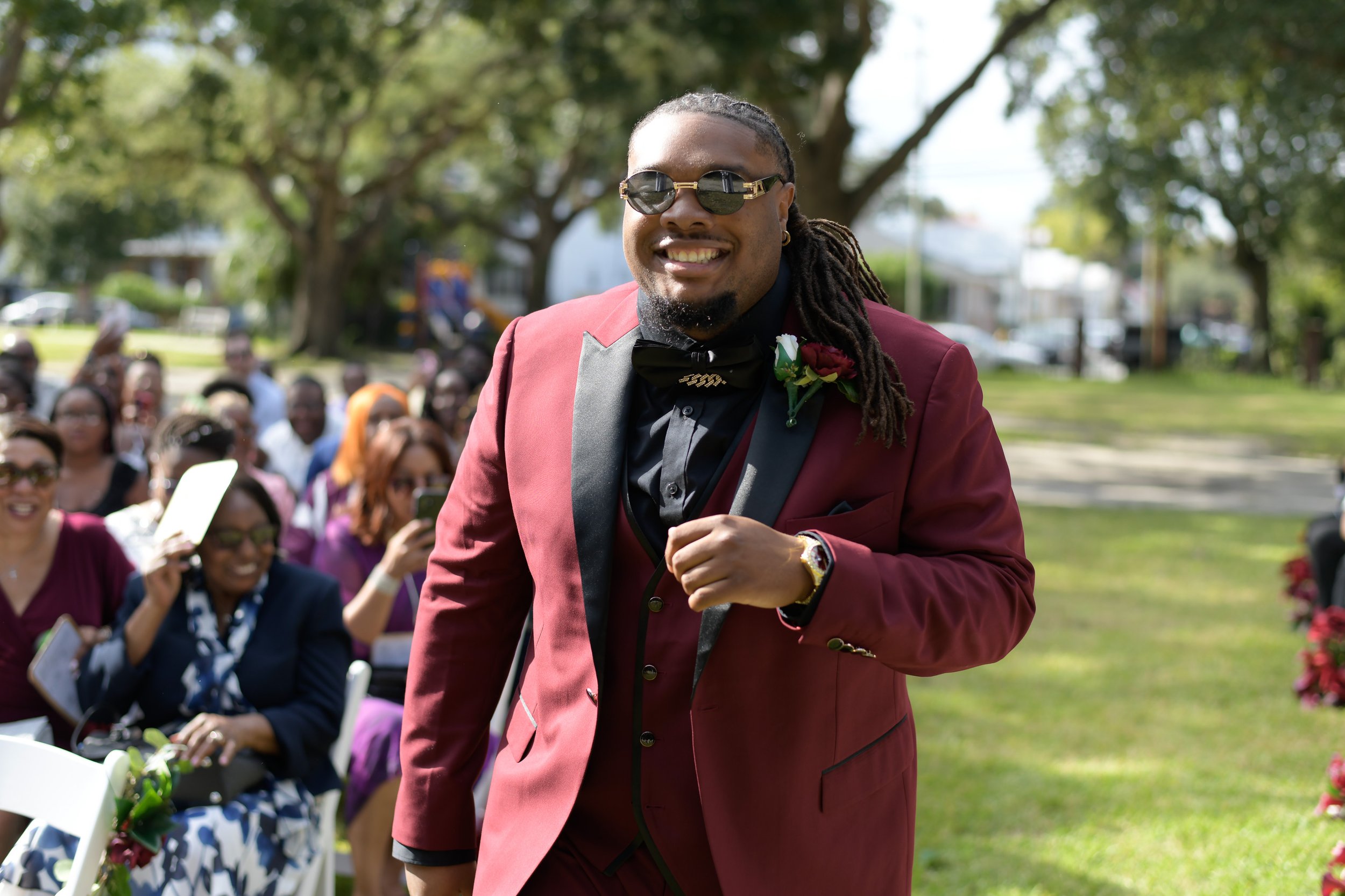 Man in a burgundy tuxedo walking outdoors at a wedding, smiling, surrounded by guests, with trees and a park in the background.