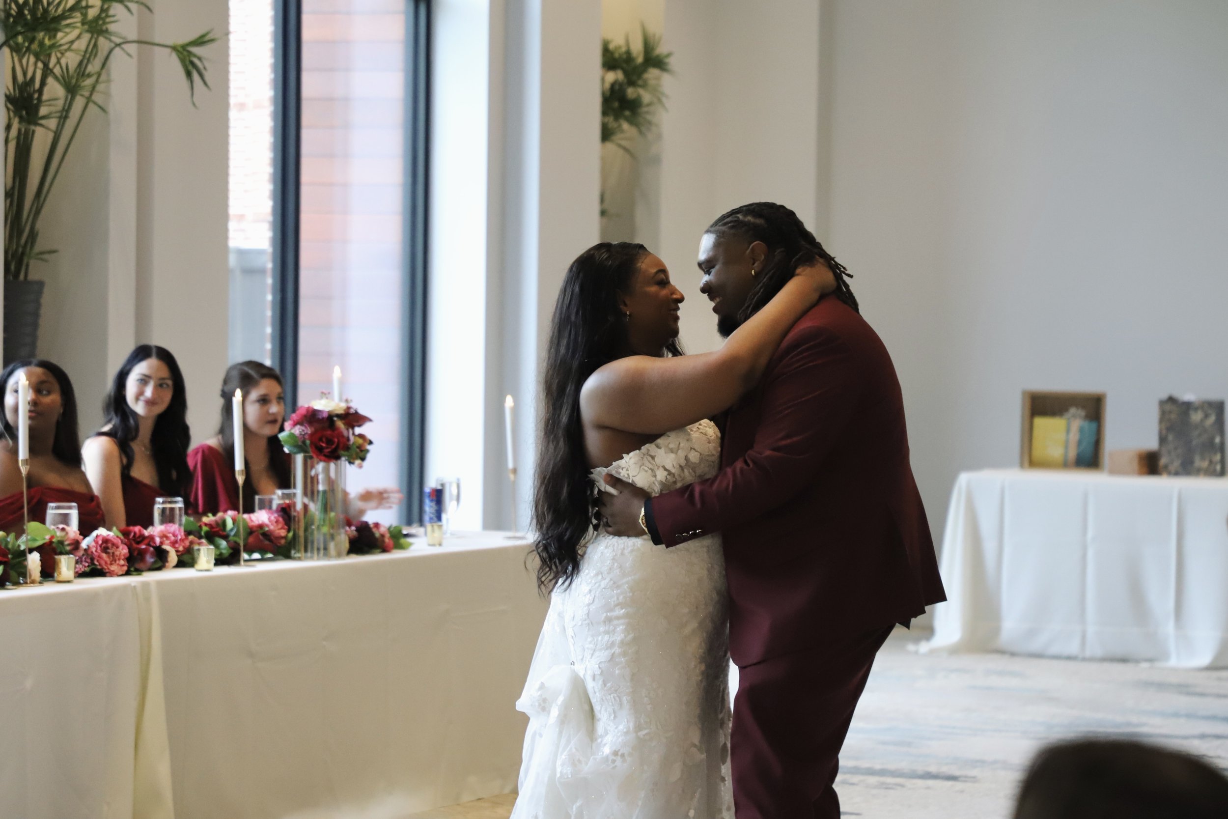 A bride and groom dancing at their wedding reception with three bridesmaids sitting at a decorated table in the background.