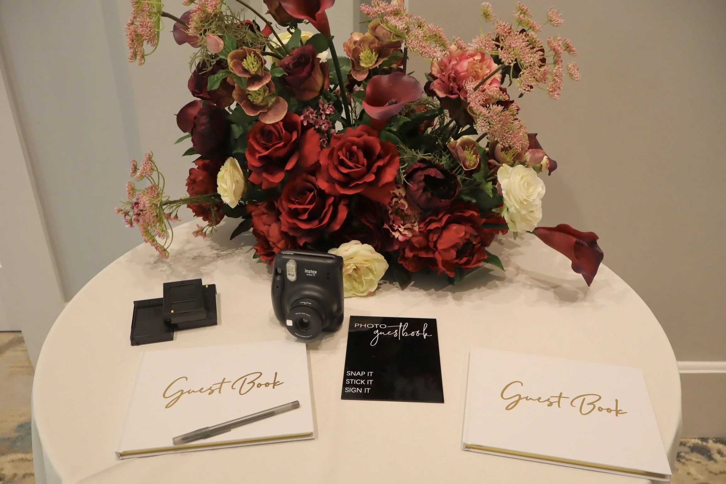 A table with a large flower arrangement of red, pink, white, and dark purple flowers, a Polaroid camera, a couple of guest books titled 'Guest Book', a pen, and a photo guest book sign with instructions to 'Snap it, Stick it, Sign it'.