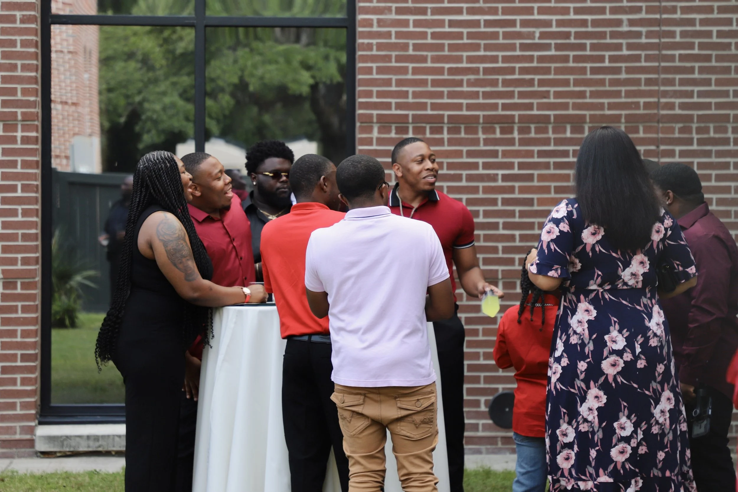 Group of people socializing outdoors near a brick building and large window, standing around a tall table, some holding drinks, wearing casual and semi-formal attire.