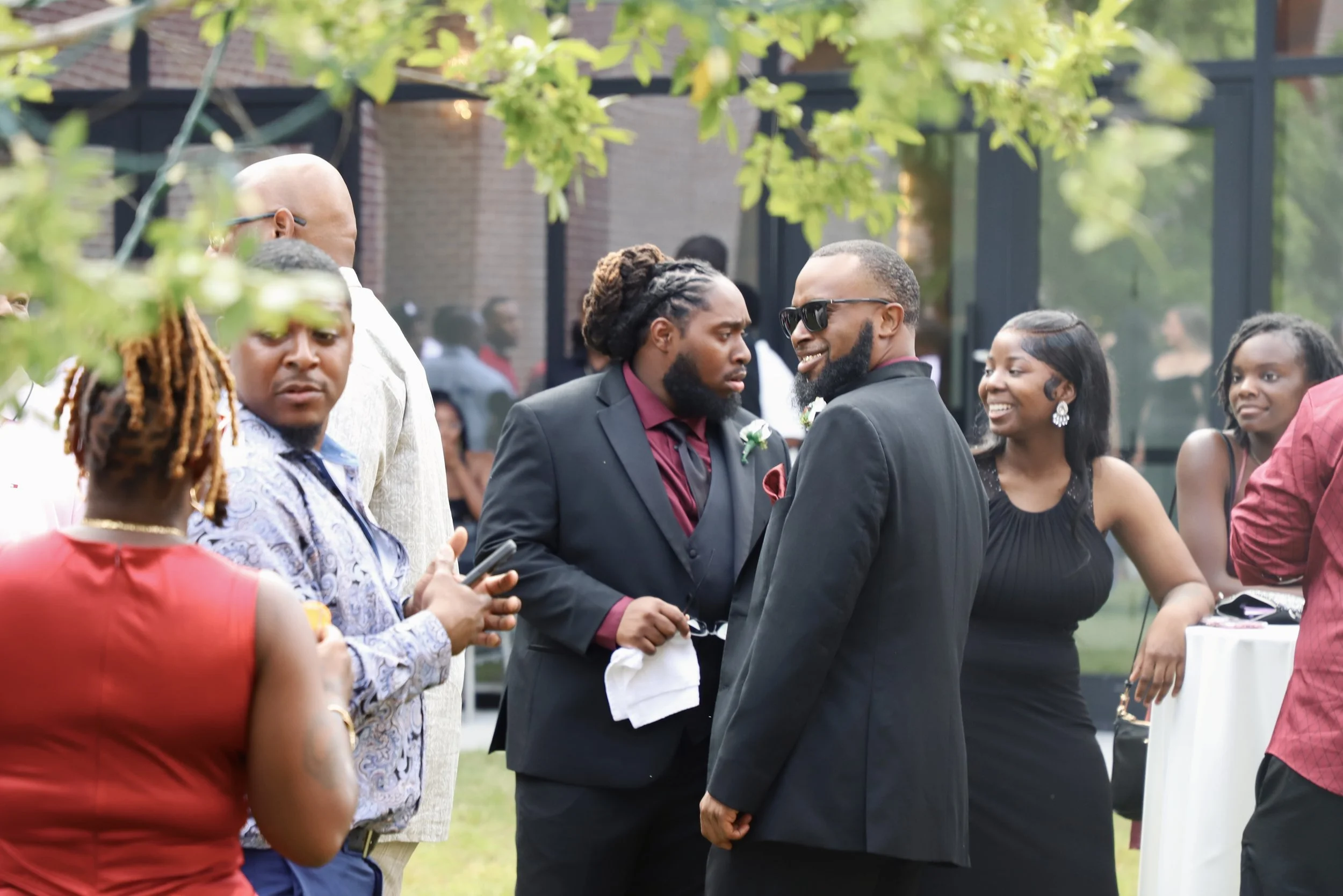 Group of people dressed in formal attire at an outdoor event, with two men in suits talking in the center, surrounded by women and men, with trees and a building in the background.