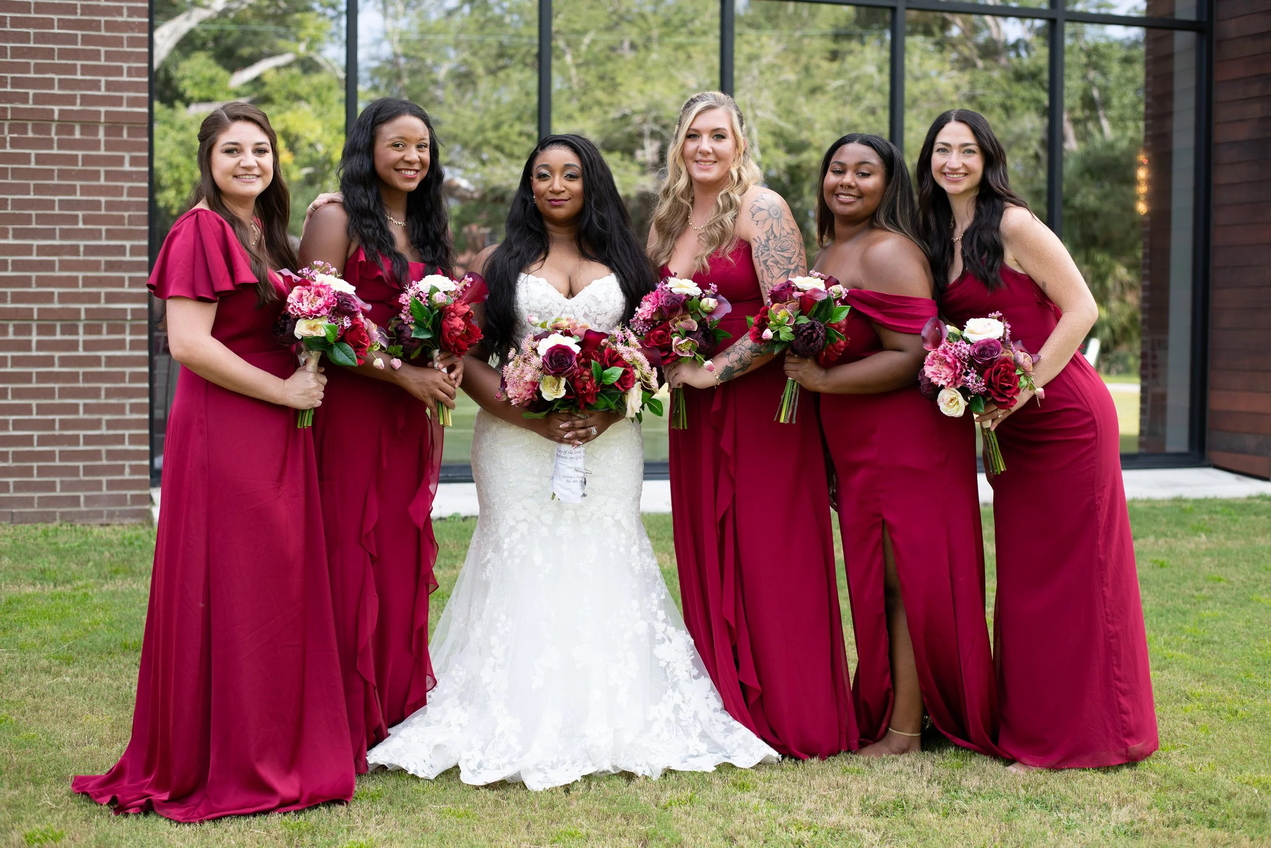 Group of women on a wedding day, including a bride in a white lace wedding dress holding a bouquet, and six bridesmaids wearing matching burgundy dresses holding bouquets, standing outside in front of a glass building with greenery in the background.