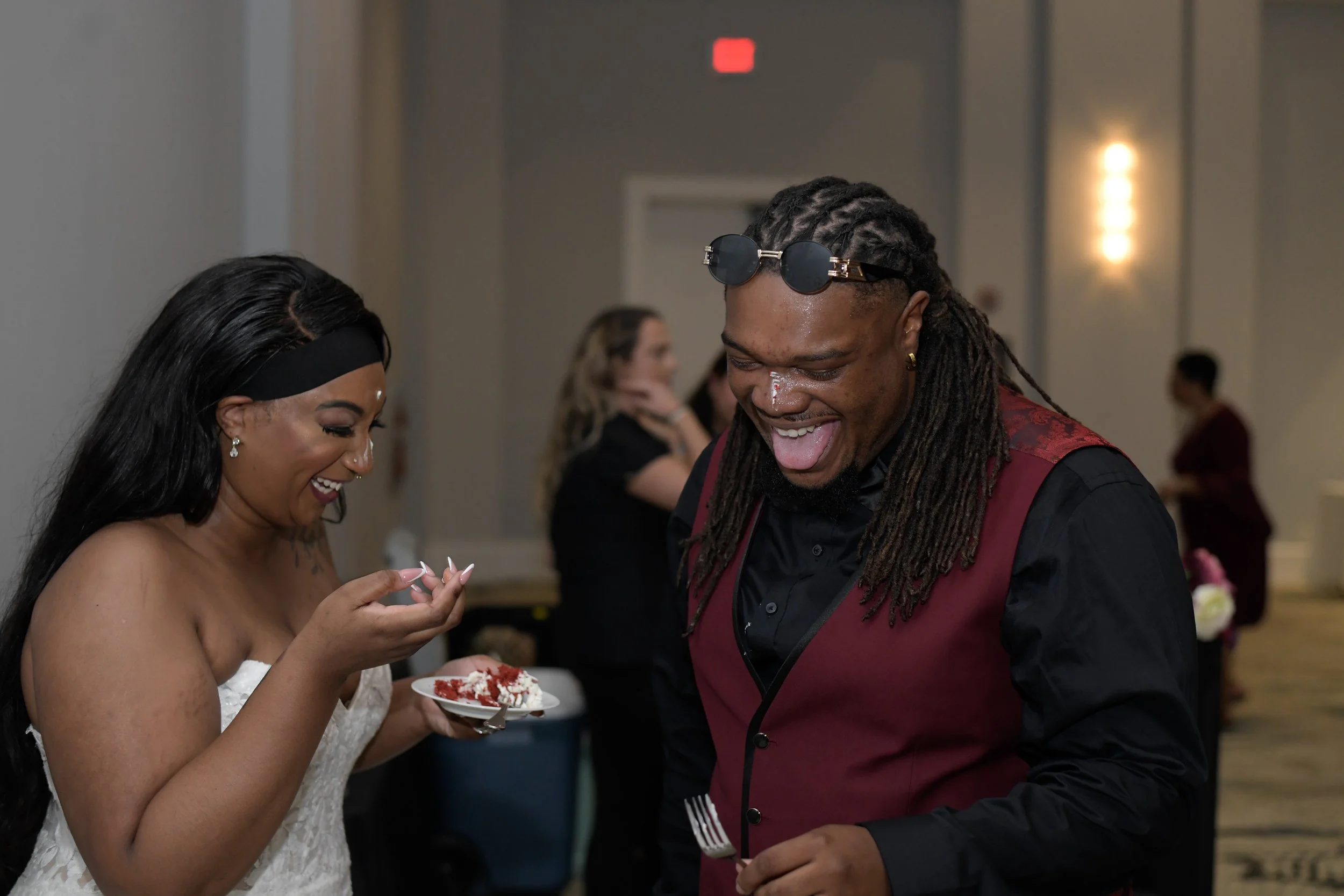A man and woman laughing and enjoying each other's company at a social event, with the woman holding a small plate of cake.