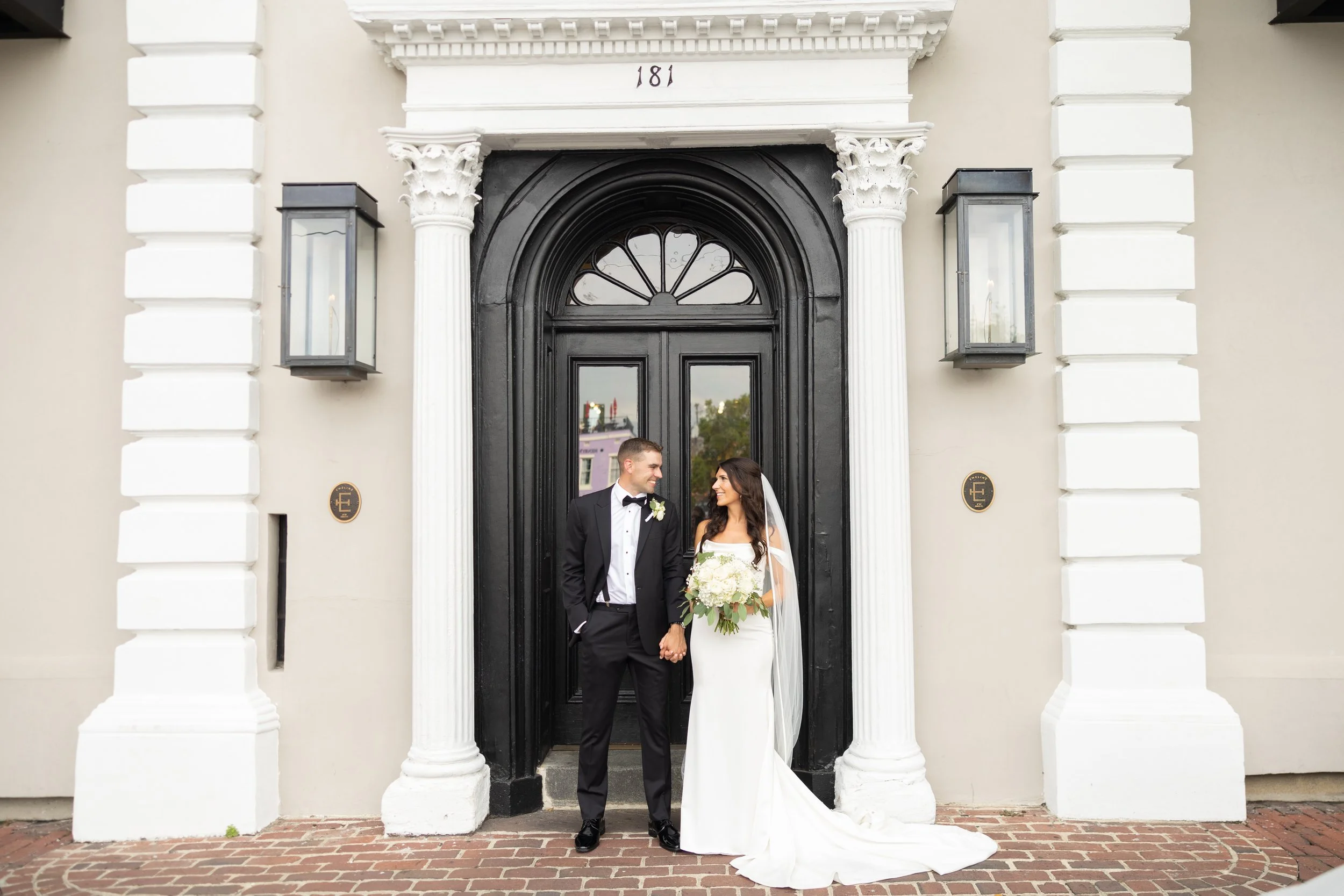 A newlywed couple standing in front of a black door with an ornate white archway. They are holding hands and looking at each other, dressed in formal wedding attire. The groom is wearing a black tuxedo and the bride a white wedding gown holding a bouquet of white flowers.