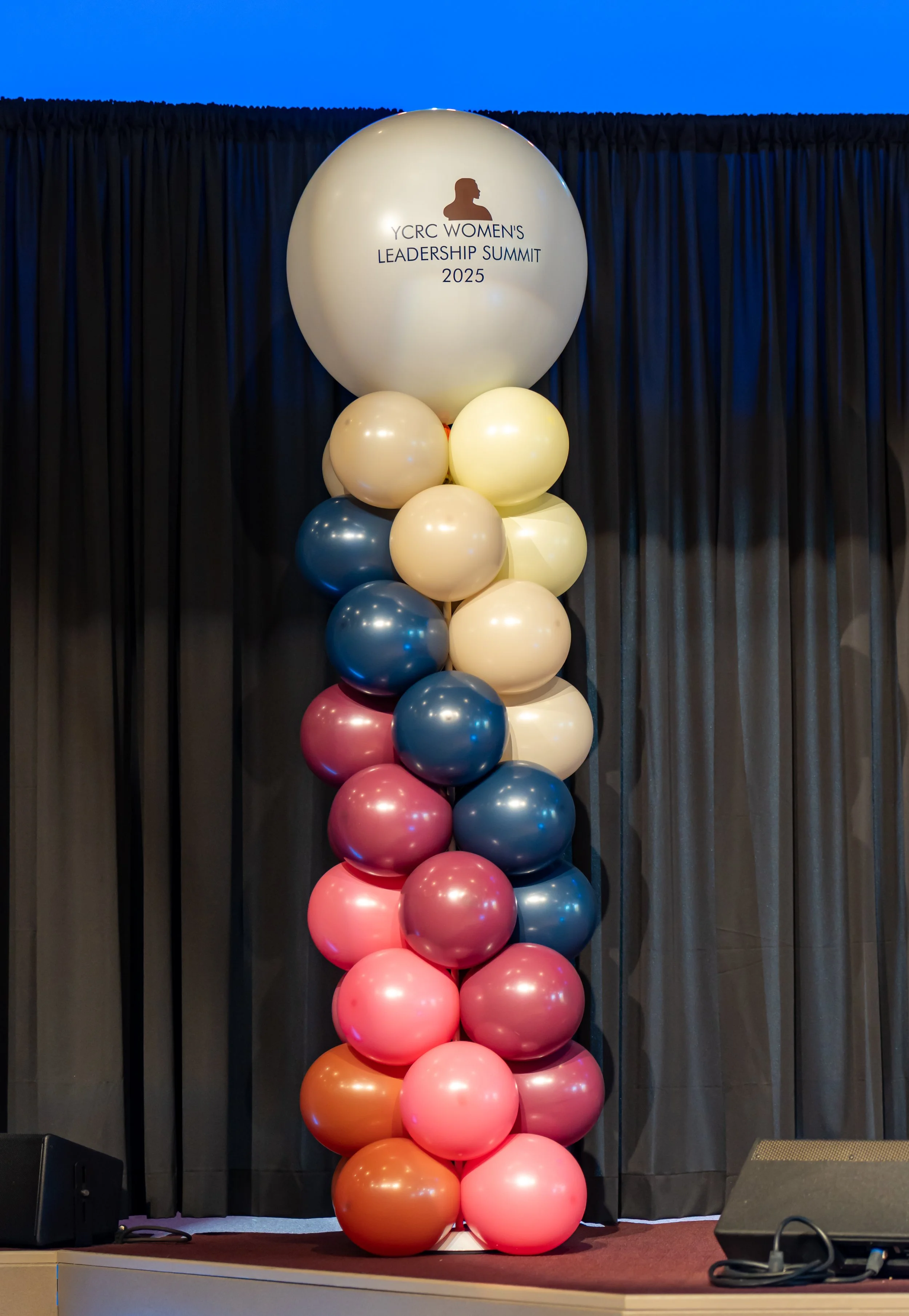 Colorful balloons arranged in a column with a large white balloon at the top displaying a logo and text for the YCRC Women's Leadership Summit 2025, set against a dark curtain backdrop.