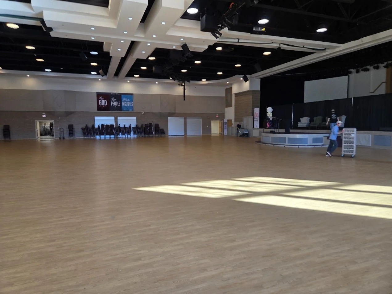 An empty auditorium or event hall with a wooden floor, a stage on the right with a person pushing a cart, and chairs stacked against the back wall. The ceiling has modern lighting and hanging speakers, with ambient light coming from windows.
