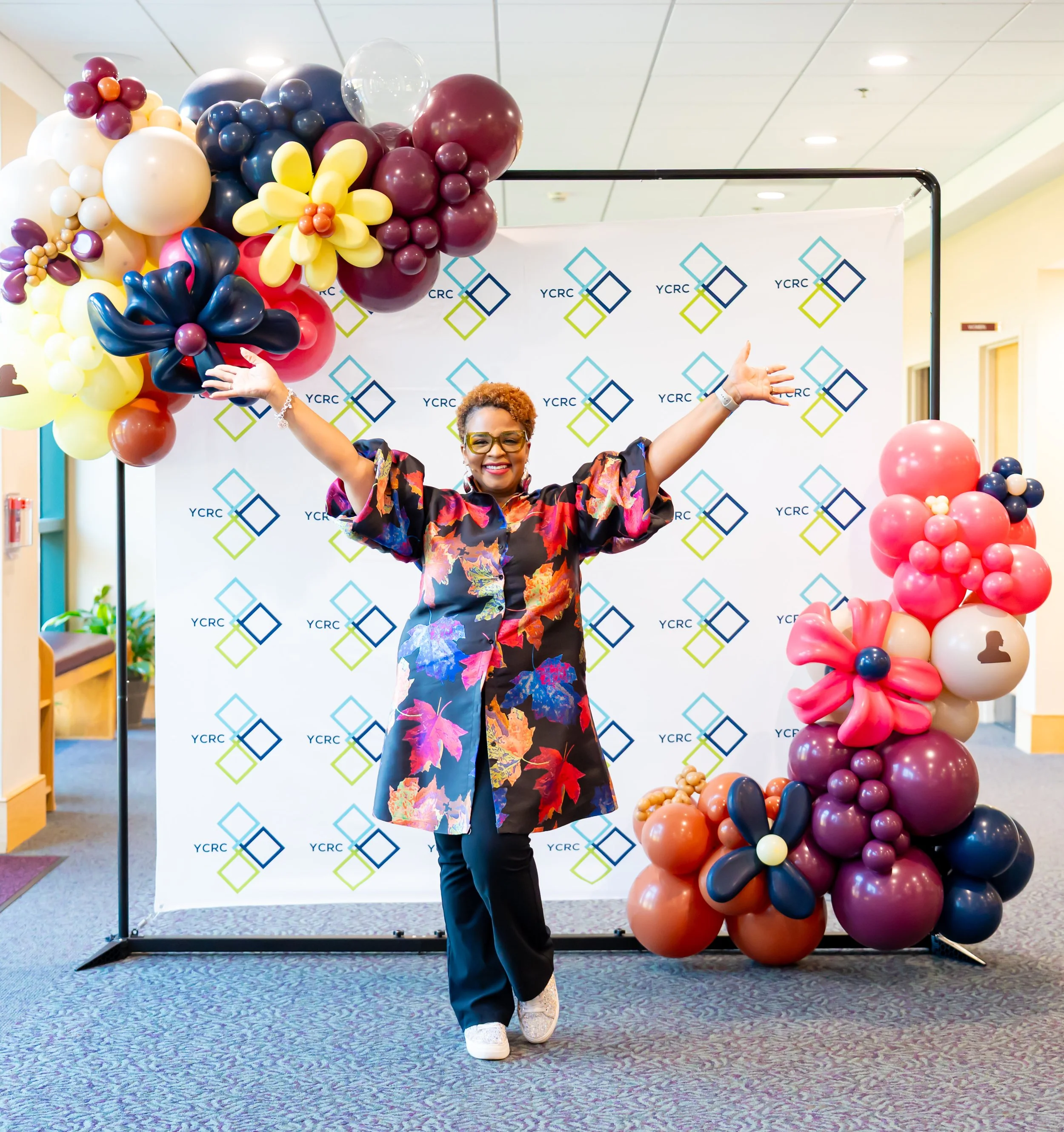 A woman with short hair and glasses wearing a floral dress with outstretched arms, standing in front of a backdrop with the YCRC logo, surrounded by colorful balloon arrangements shaped like flowers and clusters.