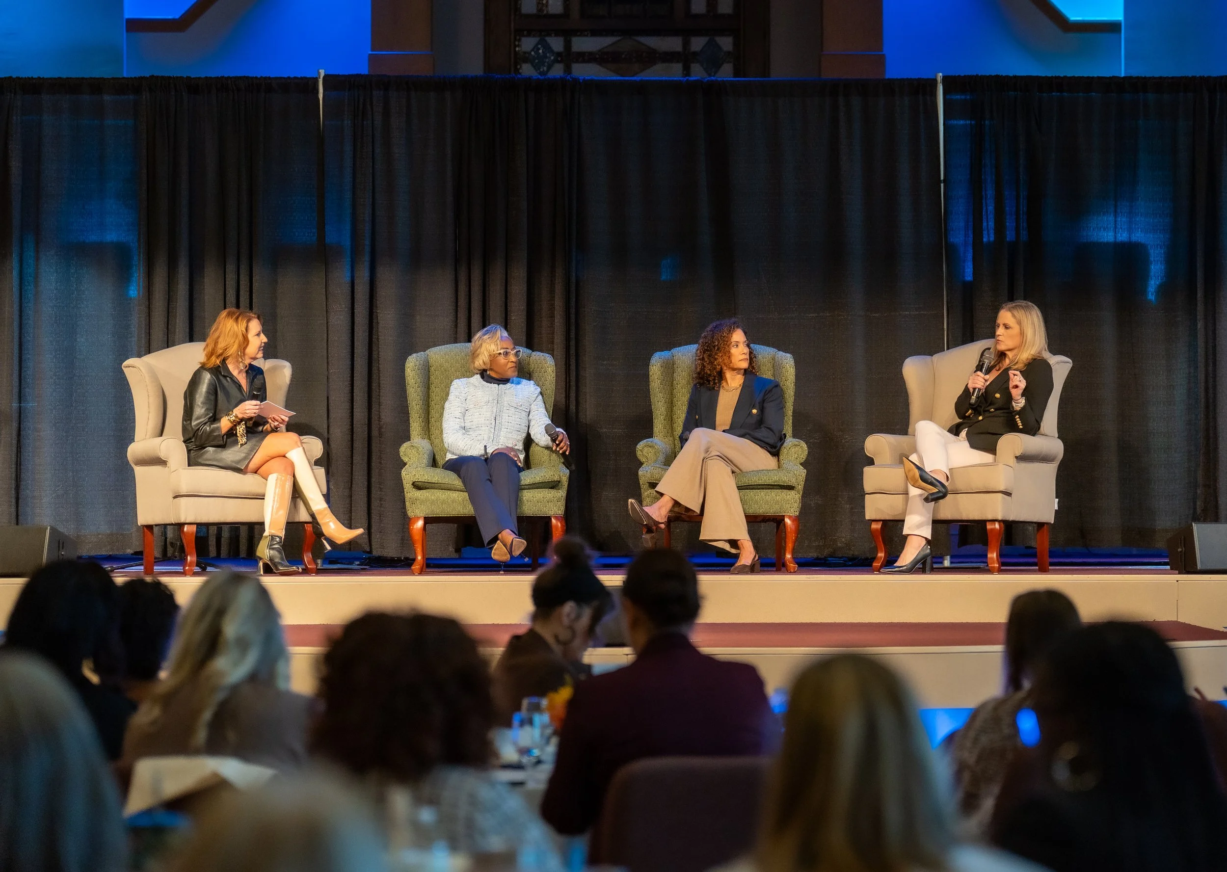 Four women sitting in chairs on a stage engaged in a discussion at a conference or panel event, with an audience watching.