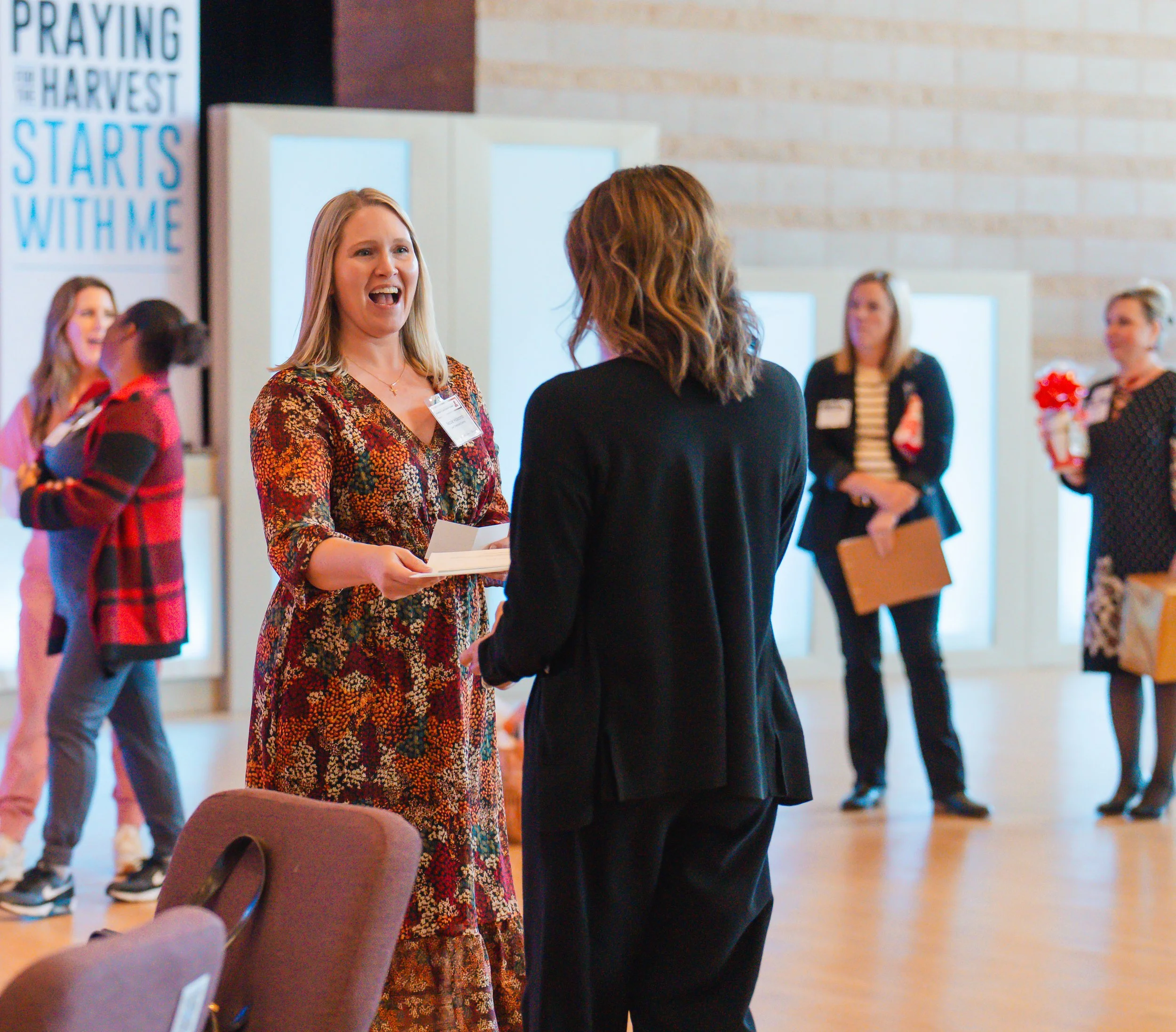 Two women are conversing at an indoor event, with one woman smiling and holding papers. Several other women are in the background, some holding flowers and a clipboard, with a banner visible that reads 'Praying the Harvest Starts with Me.'