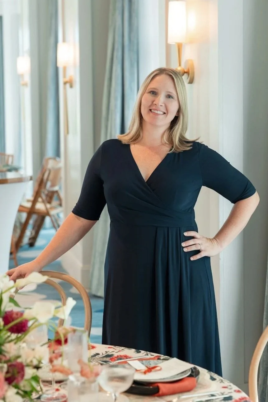 A woman with blonde hair, wearing a navy blue dress, smiling and standing in a warmly lit room with light-colored curtains and wall-mounted lamps, near a dining table with a floral centerpiece and place settings.