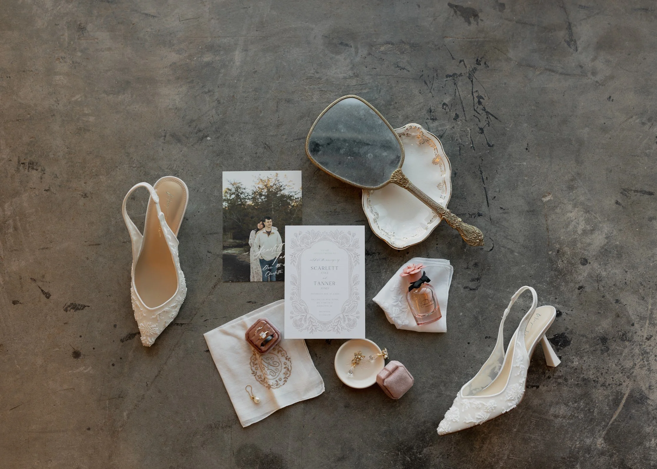 Wedding accessories on a concrete surface, including white high-heeled shoes with lace, a vintage mirror on a decorative plate, a wedding invitation, a photograph of a couple, perfume bottle, ring box, jewelry, and a small fabric pouch.
