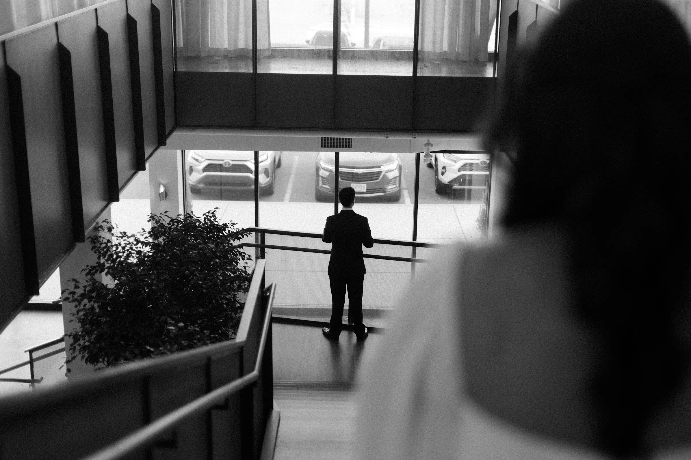 A man in a suit standing inside a building near large glass windows and looking outside at parked cars.