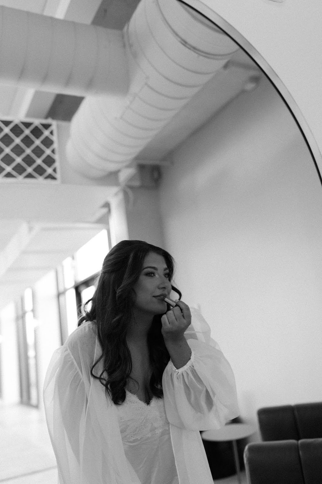 A woman with dark wavy hair applying lipstick in front of a mirror, wearing a white blouse or dress, in a room with modern decor and large windows.