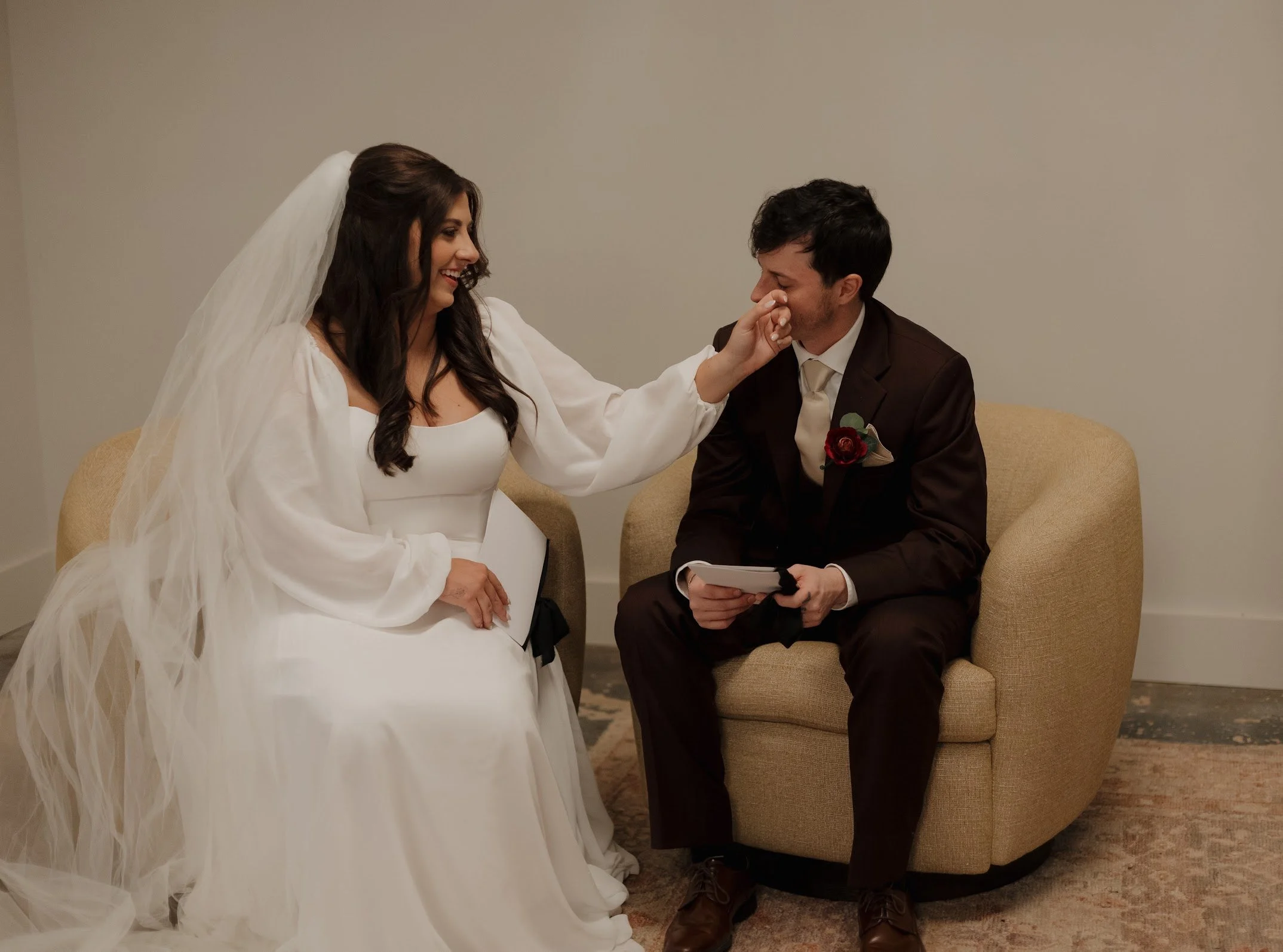 A bride and groom sitting on a beige armchair, with the bride wiping tears from the groom's face during their wedding ceremony.