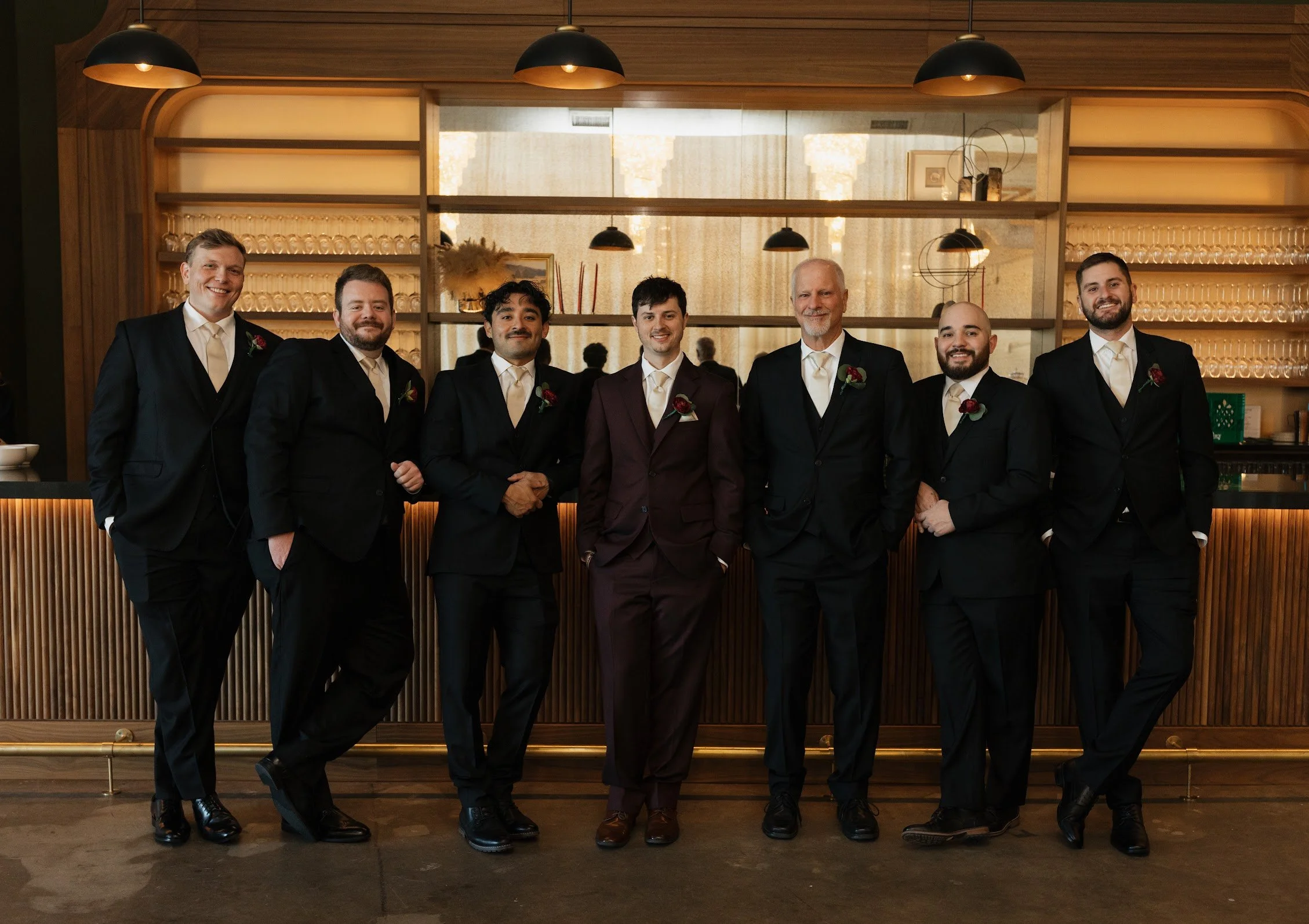 A group of seven men in tuxedos standing side by side in a modern, warmly lit bar or restaurant setting, smiling at the camera.