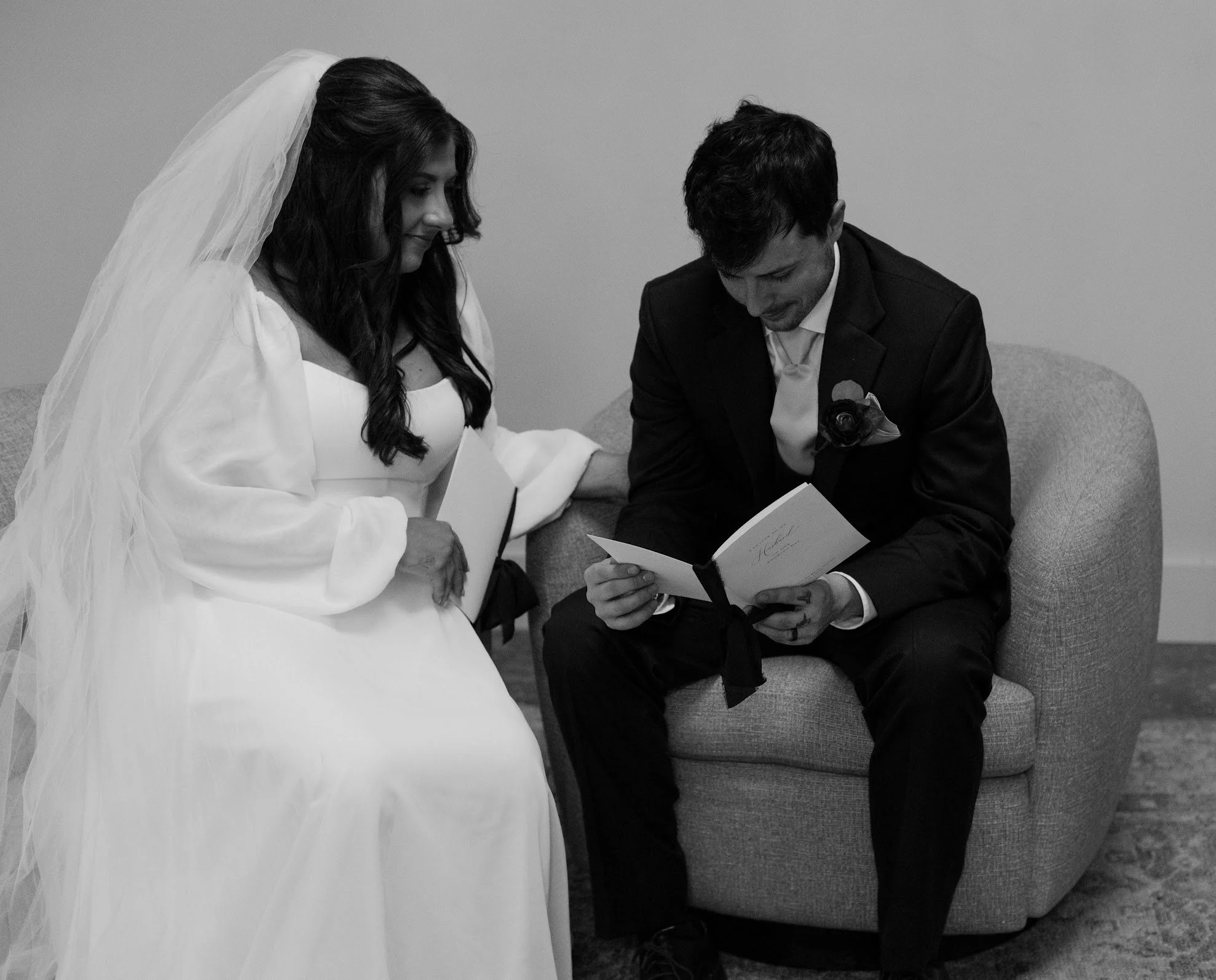 Black and white photo of a bride in a wedding dress and veil sitting on a couch beside a groom in a suit reading a card.