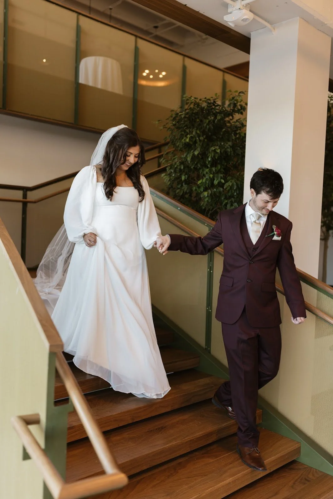 A bride and groom holding hands as they descend a wooden staircase during their wedding celebration indoors.
