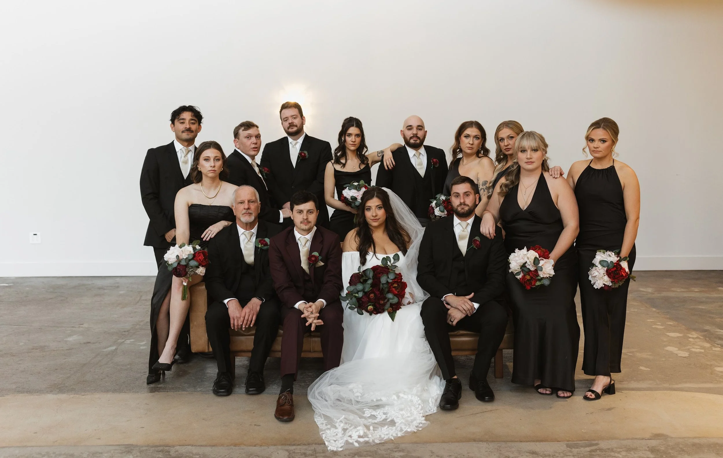 A large wedding party poses together indoors against a plain white wall. The bride in a white gown with lace details holds a bouquet, seated in the center. The groom in a dark suit with a white shirt and boutonniere sits beside her. Bridesmaids and g