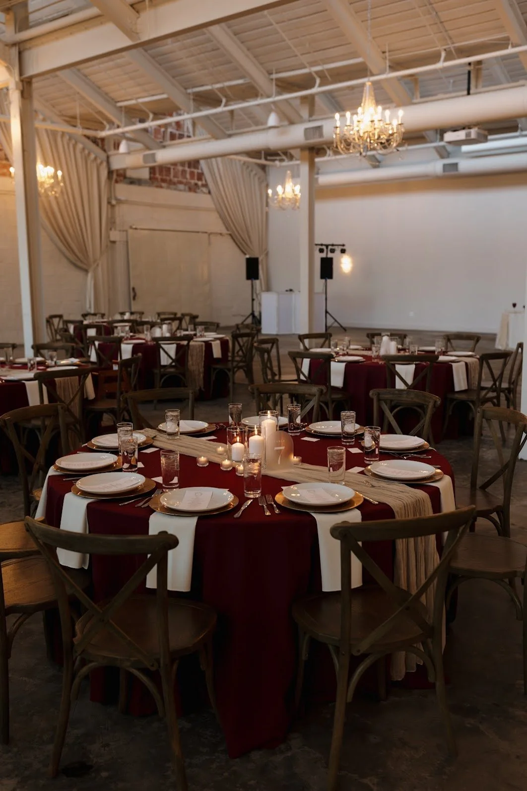 Elegant banquet hall set for a celebration with tables covered in burgundy and white tablecloths, candle centerpieces, plates, glasses, and cutlery, decorated with a beige table runner and candles.