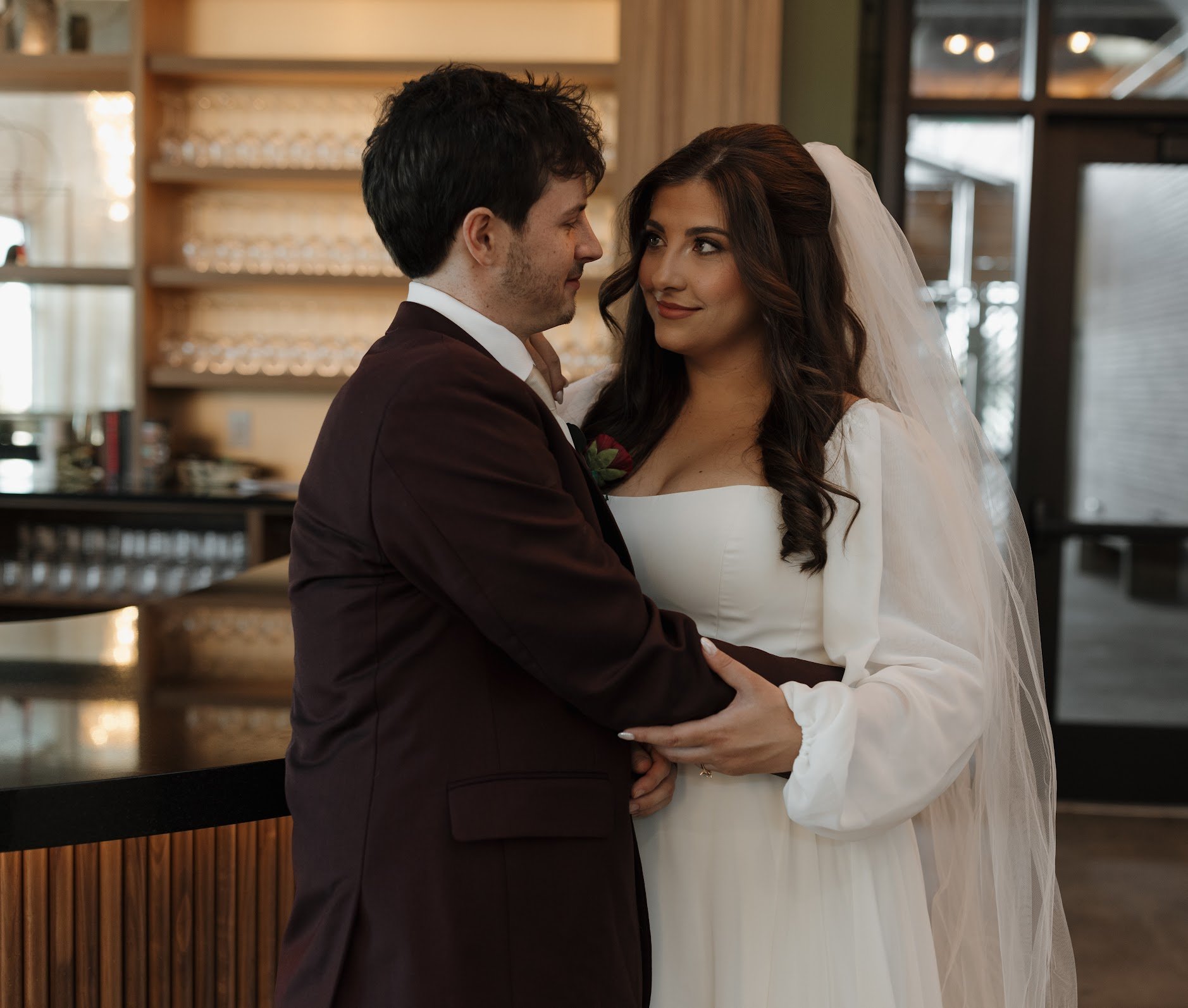 A bride and groom sharing a moment, holding hands and looking at each other warmly in a rustic indoor setting.