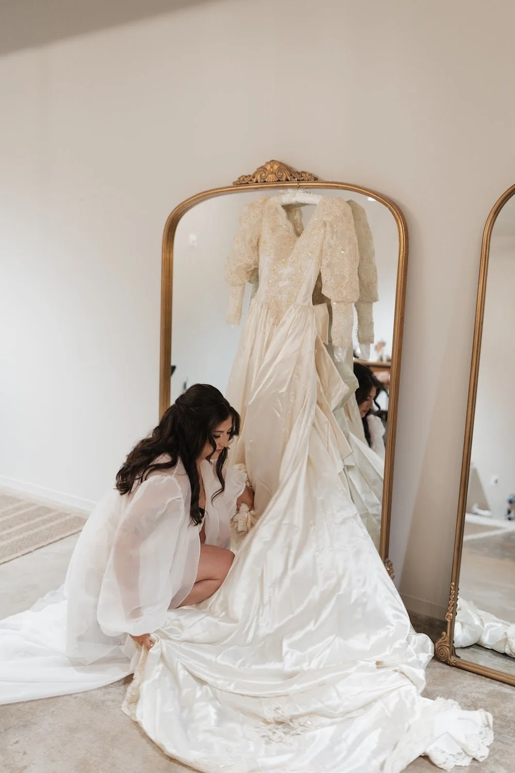 A woman in white, likely a bride, kneeling to adjust a wedding gown in front of a large ornate mirror.