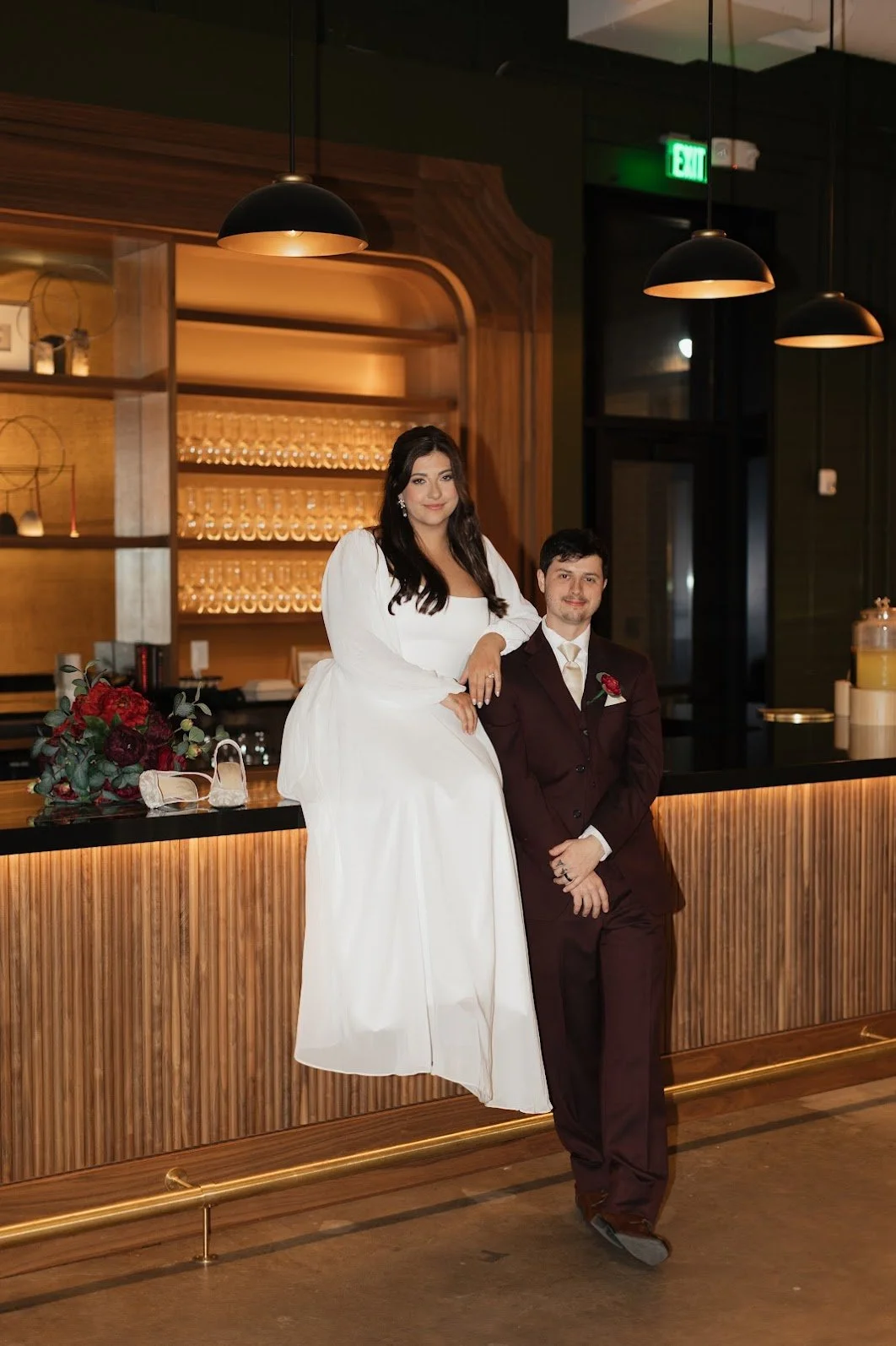 A woman in a white wedding dress sitting on a bar counter, with a man in a dark maroon suit standing beside her, inside a modern venue with warm lighting and glassware on shelves.