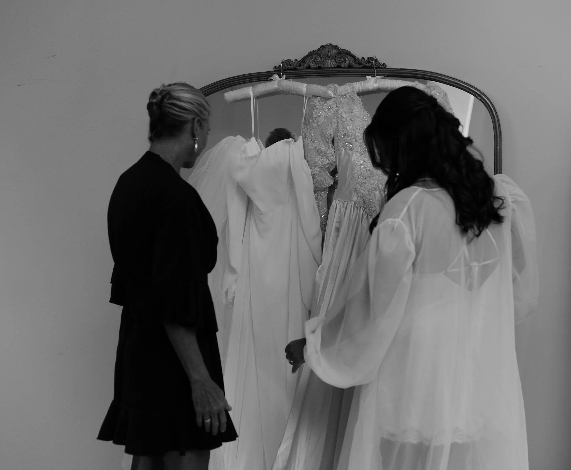 Two women looking at dresses on a clothing rack, with one wearing a dark dress and the other a light, sheer dress.