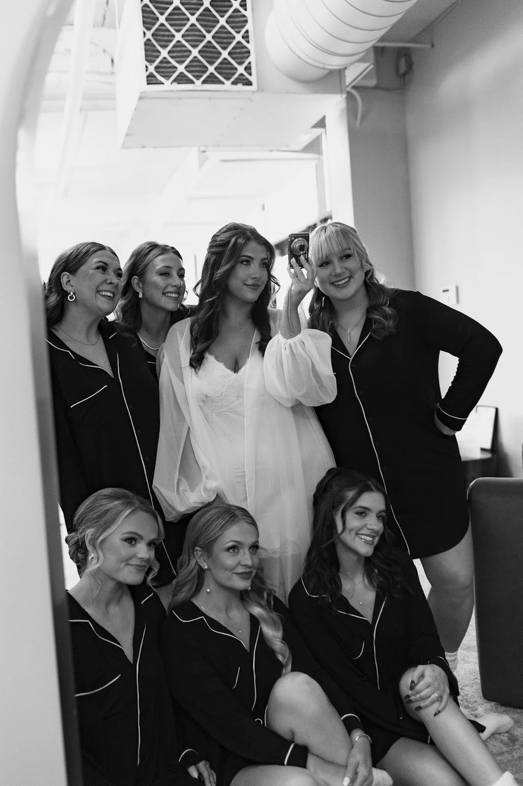 A group of women, in pajamas, taking a mirror selfie in a room. One woman is wearing a white robe, and others are dressed in black pajamas with white piping.