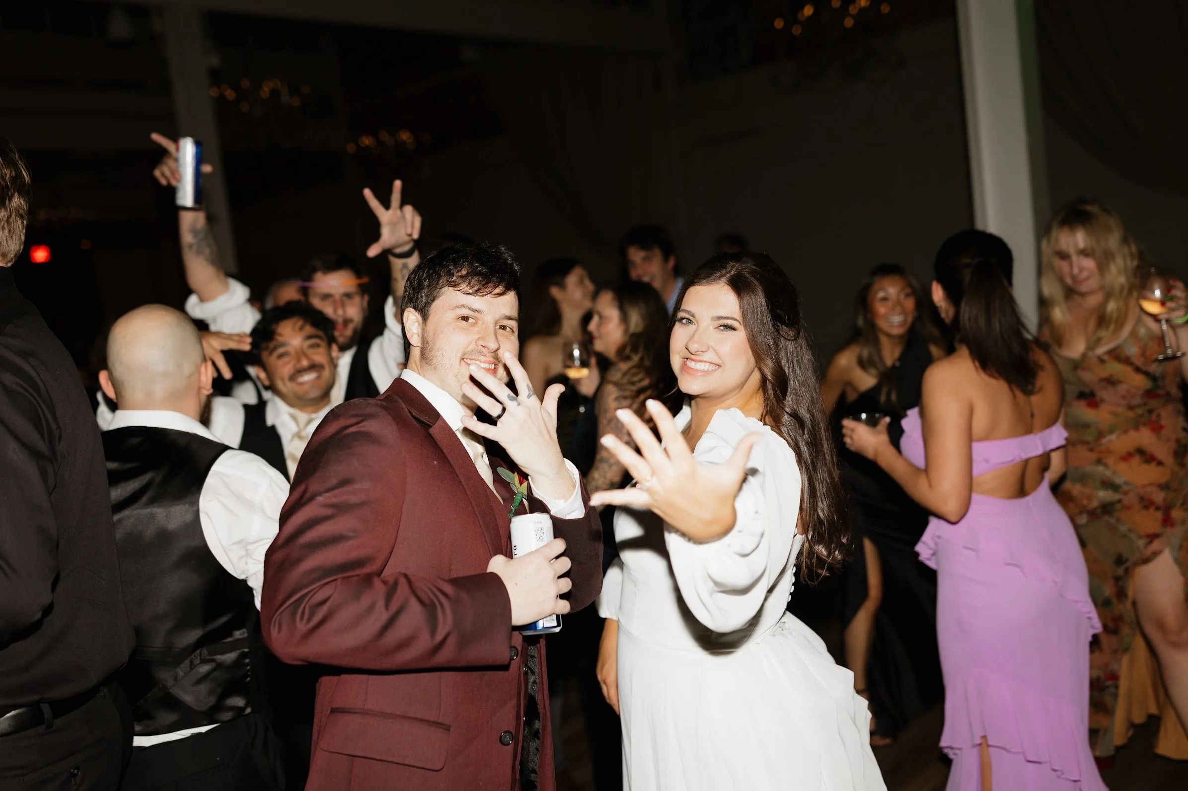 A group of people at a party or celebration dancing and smiling. Prominently, a man in a burgundy suit and woman in a white dress are in the foreground showing playful gestures and smiling. In the background, others are enjoying the event with drinks