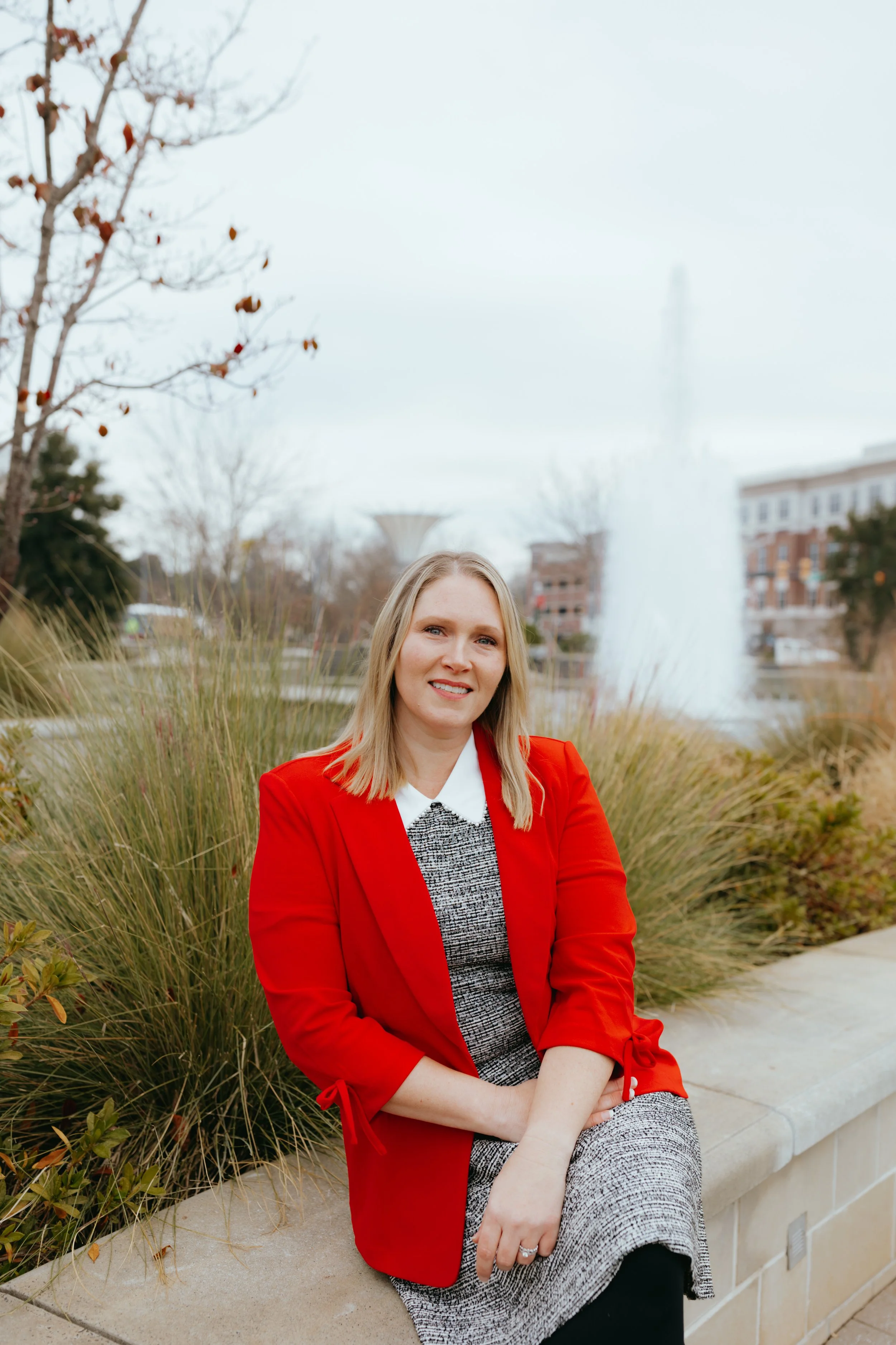 A woman sitting on a concrete bench near tall grass and trees, wearing a red blazer over a gray and white dress, with a park and fountain in the background on an overcast day.