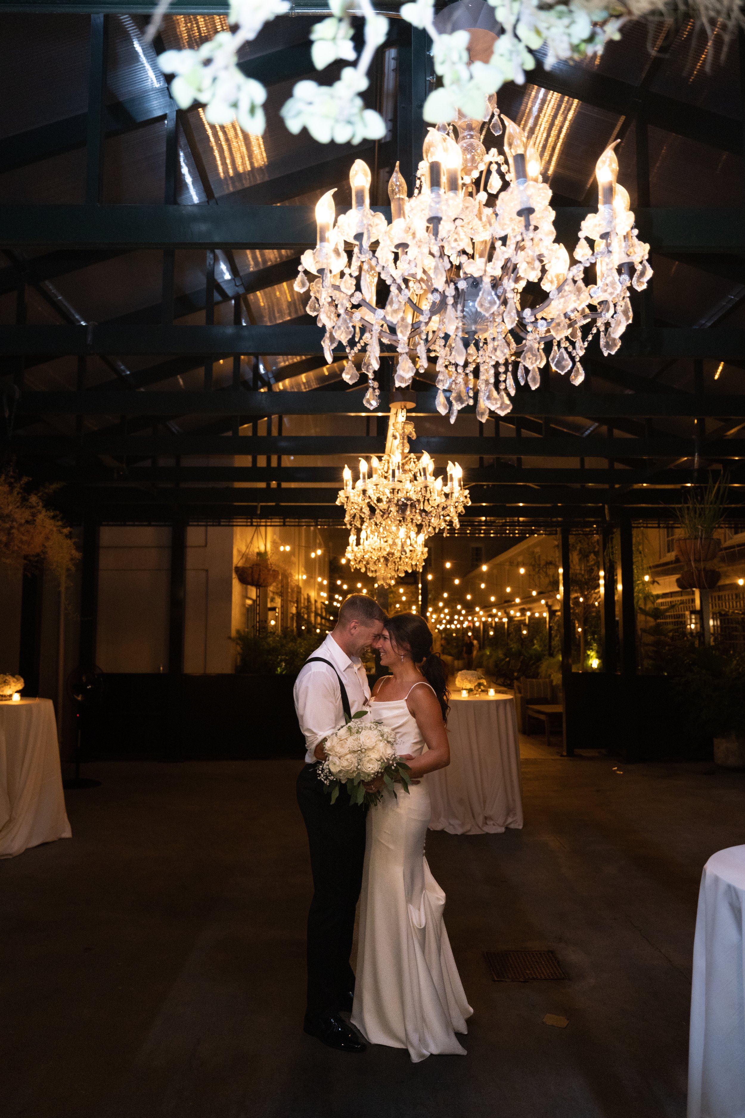 A bride and groom standing close together and smiling at each other under a chandelier during a wedding reception at night.