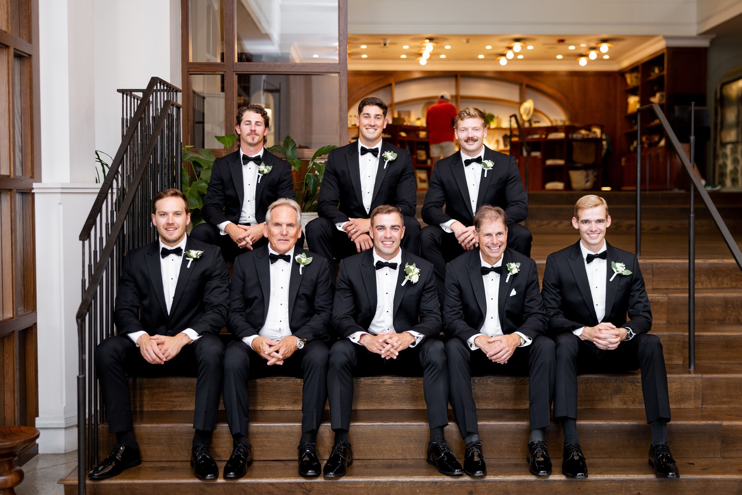 A group of ten men in tuxedos sitting and standing on wooden stairs inside a building, likely at a wedding celebration.