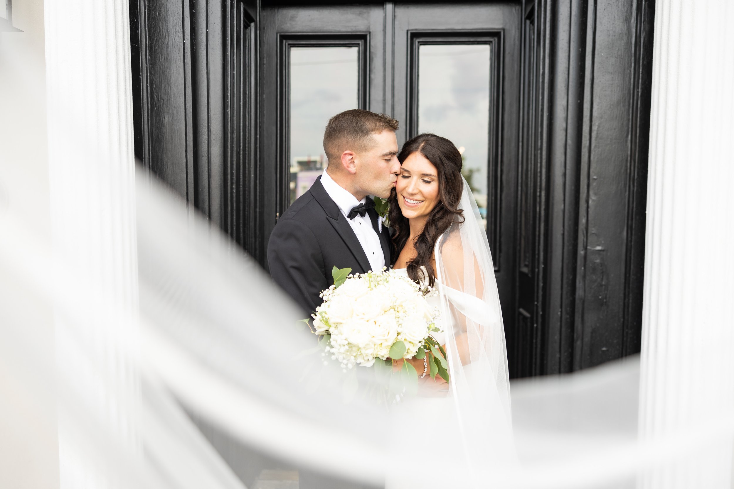 A newlywed couple shares a kiss on their wedding day. The groom is in a black tuxedo with a bow tie, and the bride is in a white wedding dress holding a bouquet of white flowers. They are standing in front of a black door, smiling happily.