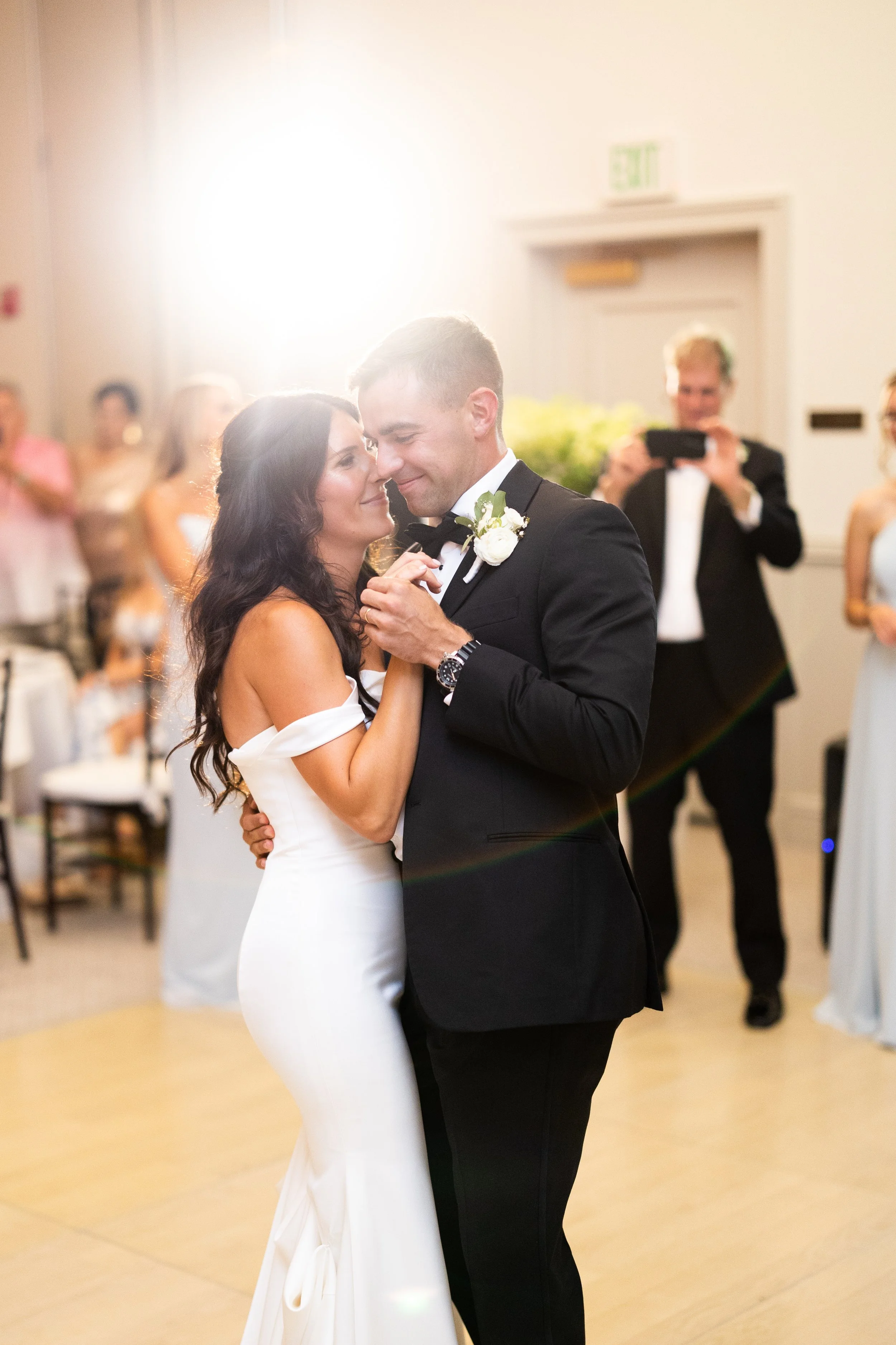 A bride and groom share a dance at their wedding reception. The bride is in a white gown with off-the-shoulder sleeves, and the groom is in a black tuxedo. Guests are watching in the background, and a person is taking a photo.