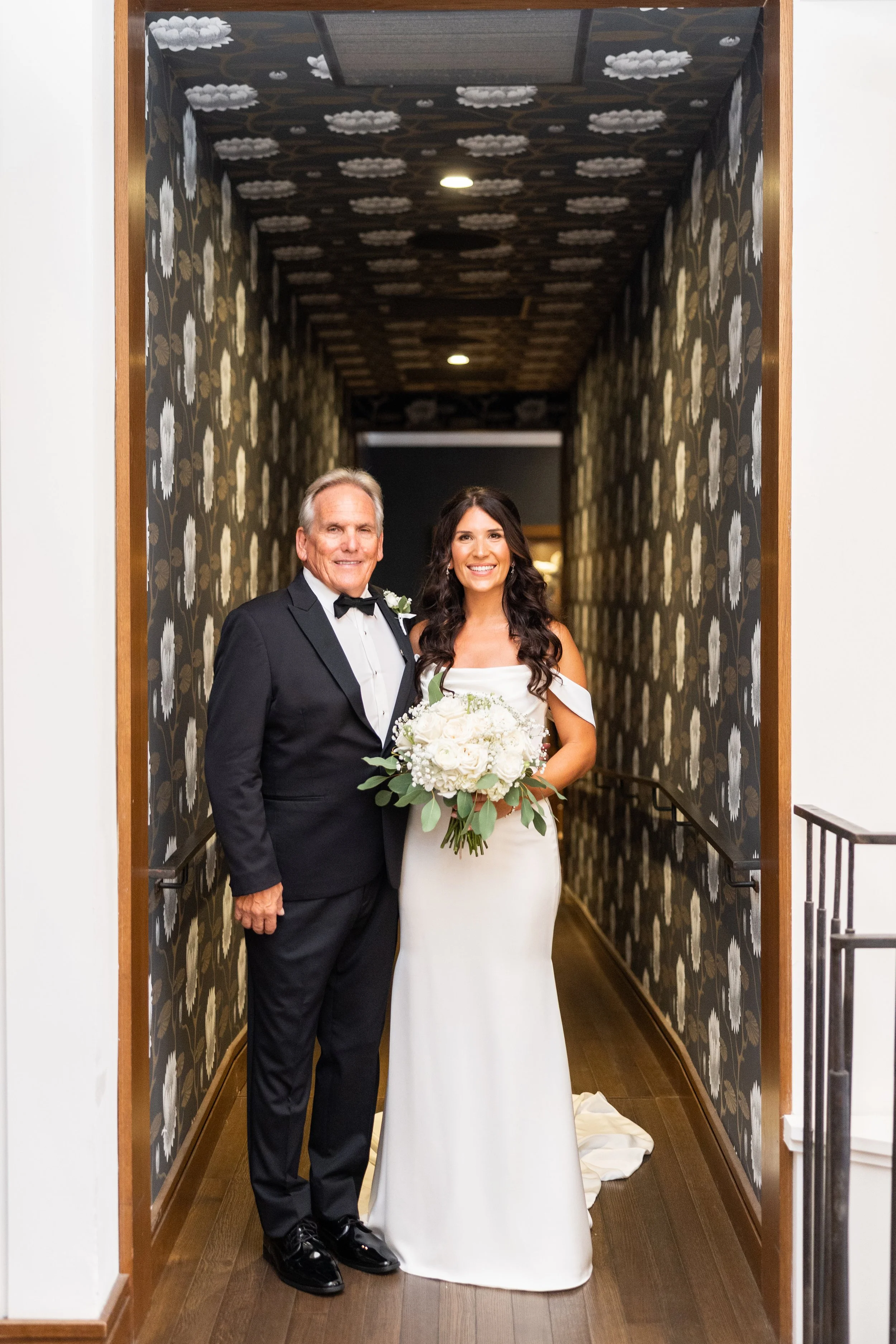 A bride and an older man, likely her father, standing in a decorated hallway. The bride is holding a bouquet of white flowers and wearing a white wedding dress. The older man is wearing a black tuxedo with a bow tie. They are smiling at the camera.