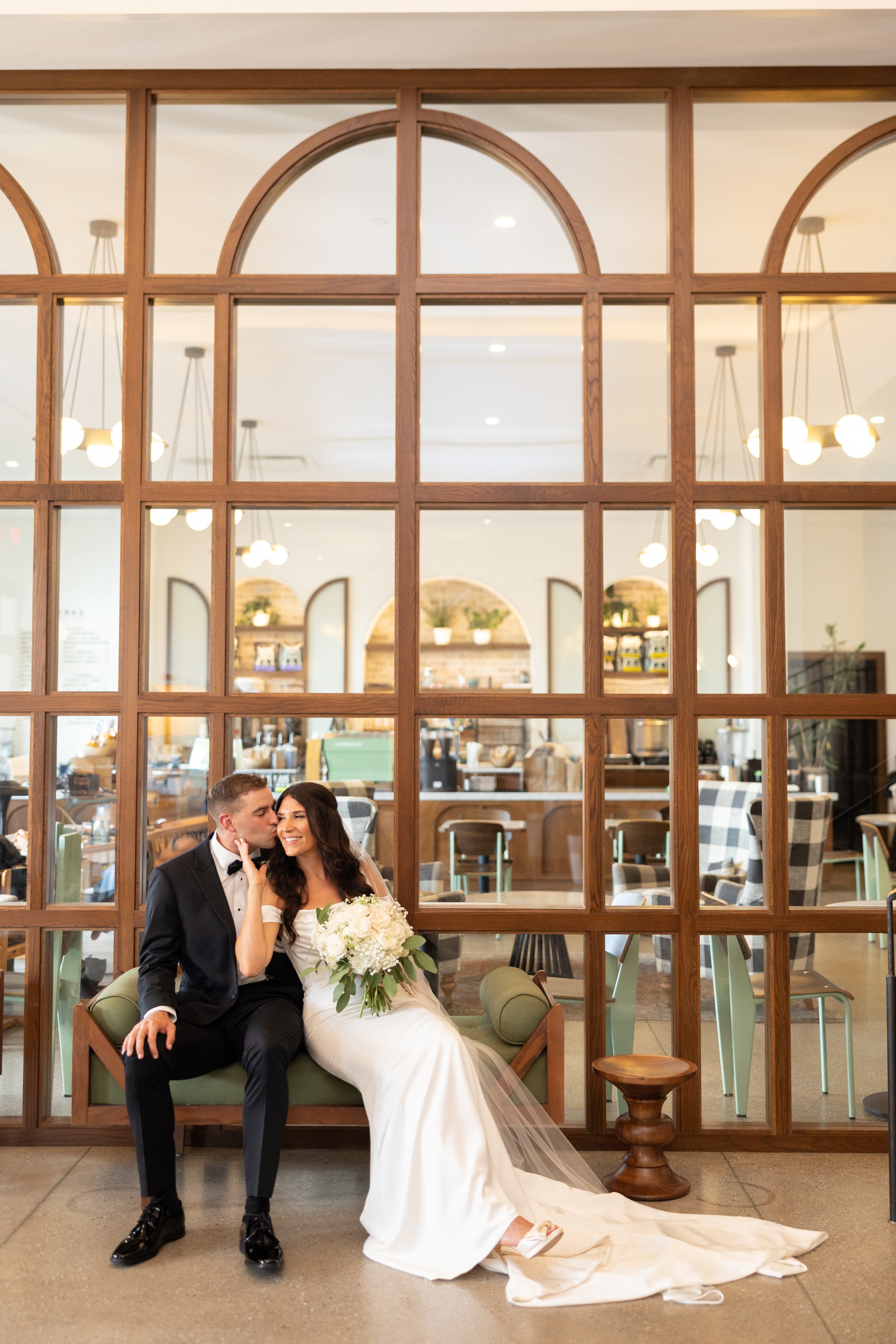A bride and groom sitting on a sofa in a restaurant, with a wooden lattice divider behind them. The bride is holding a bouquet of white flowers and smiling as the groom kisses her cheek.