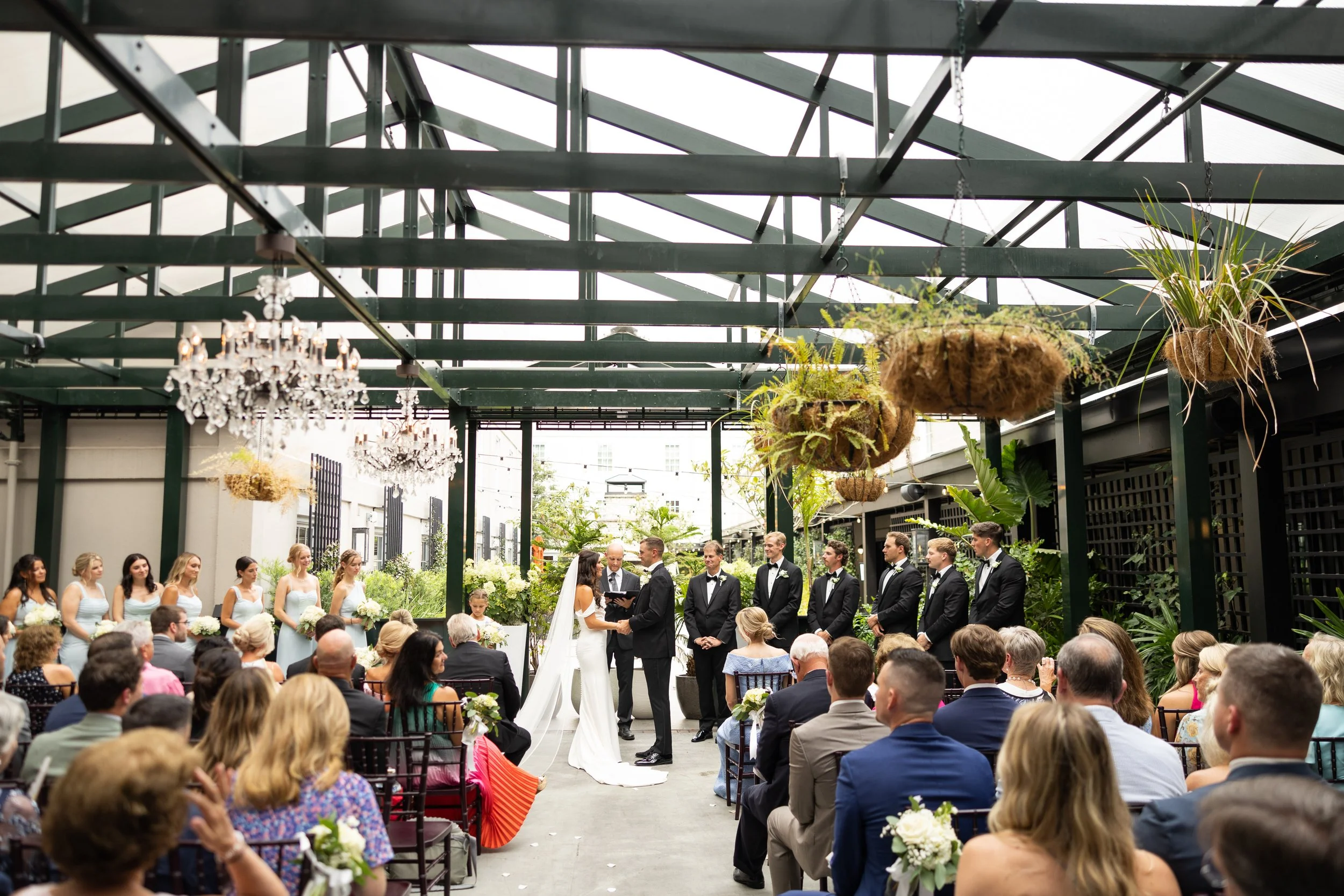 A wedding ceremony taking place indoors with guests seated facing a bride and groom, officiant, and bridal party, in a space decorated with hanging plants and chandeliers.