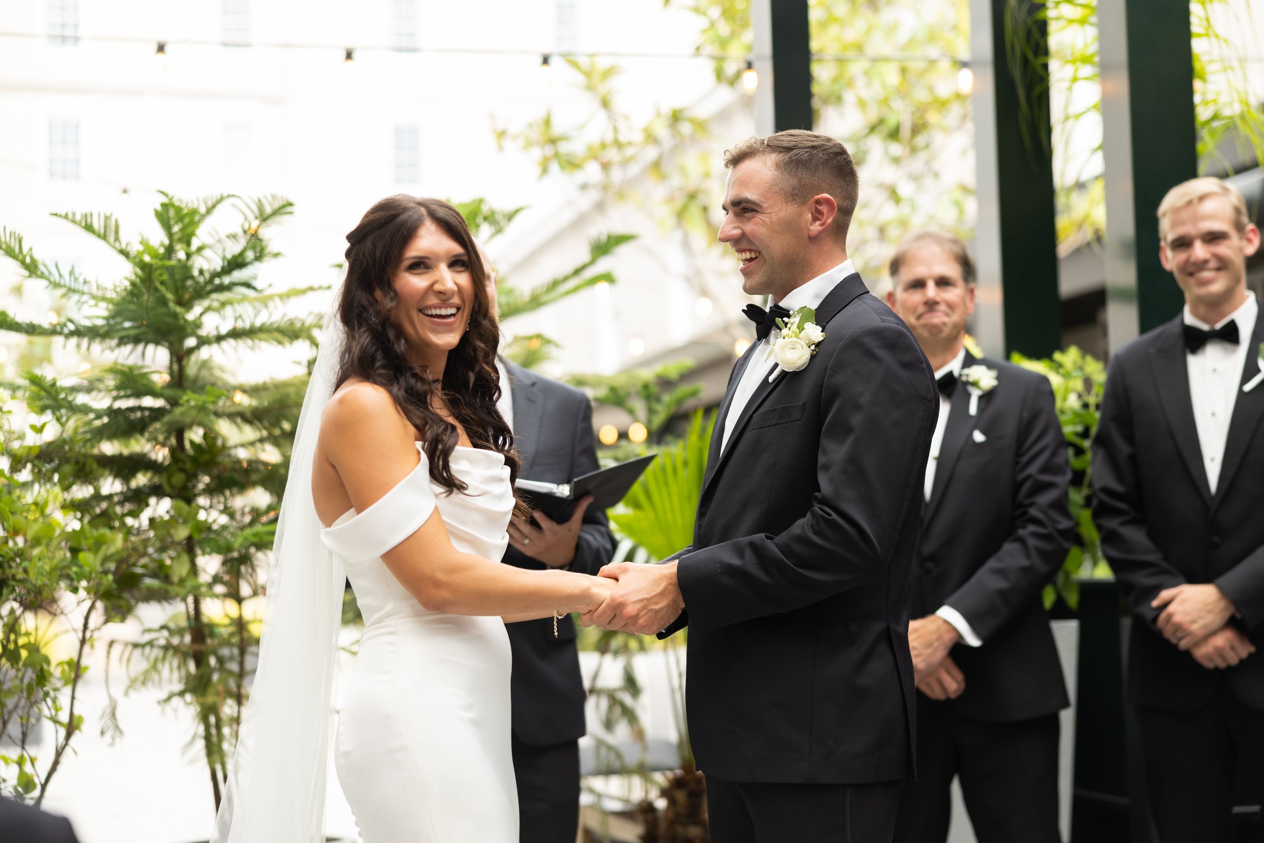 A bride and groom exchanging wedding vows in a greenhouse, surrounded by two groomsmen, with lush greenery and sunlight in the background.