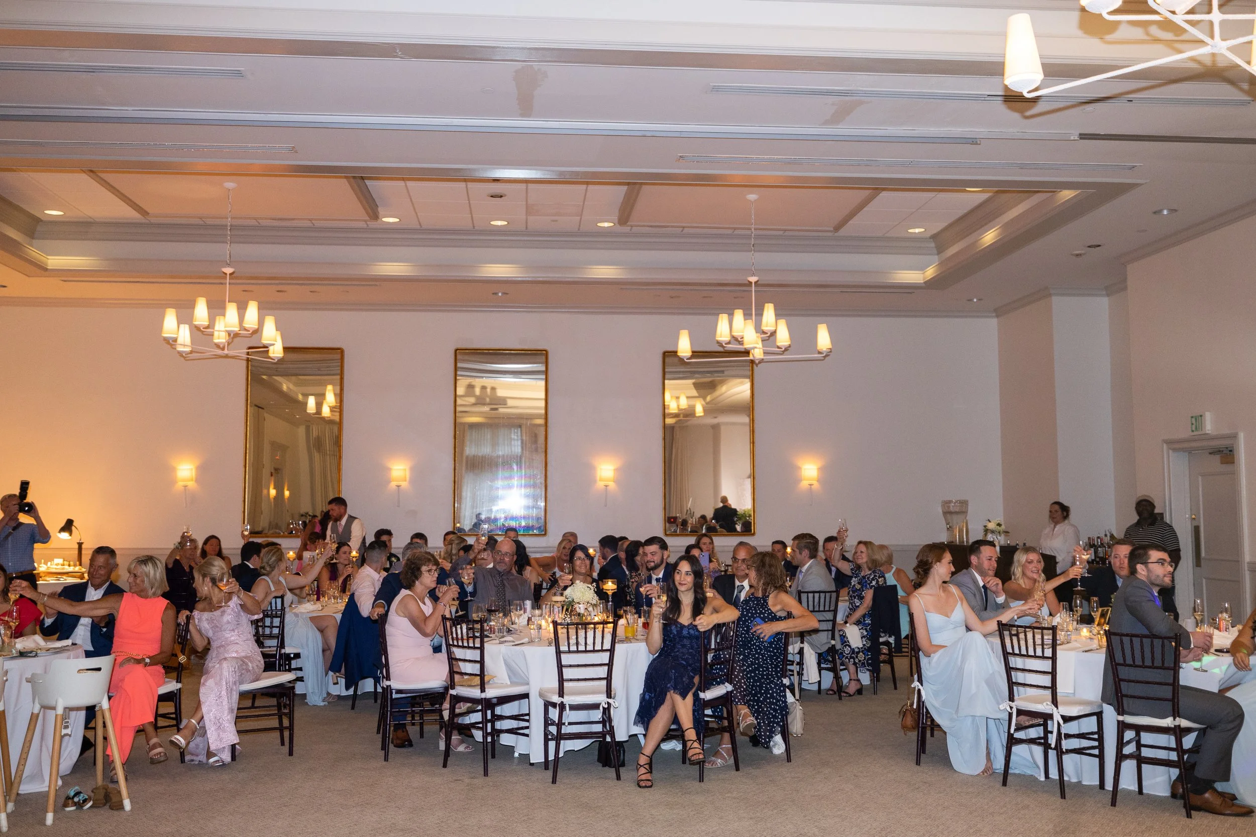People at a formal event dinner in a banquet hall with mirrors on the wall, chandeliers, and tables decorated with flowers and candles, raising glasses for a toast.