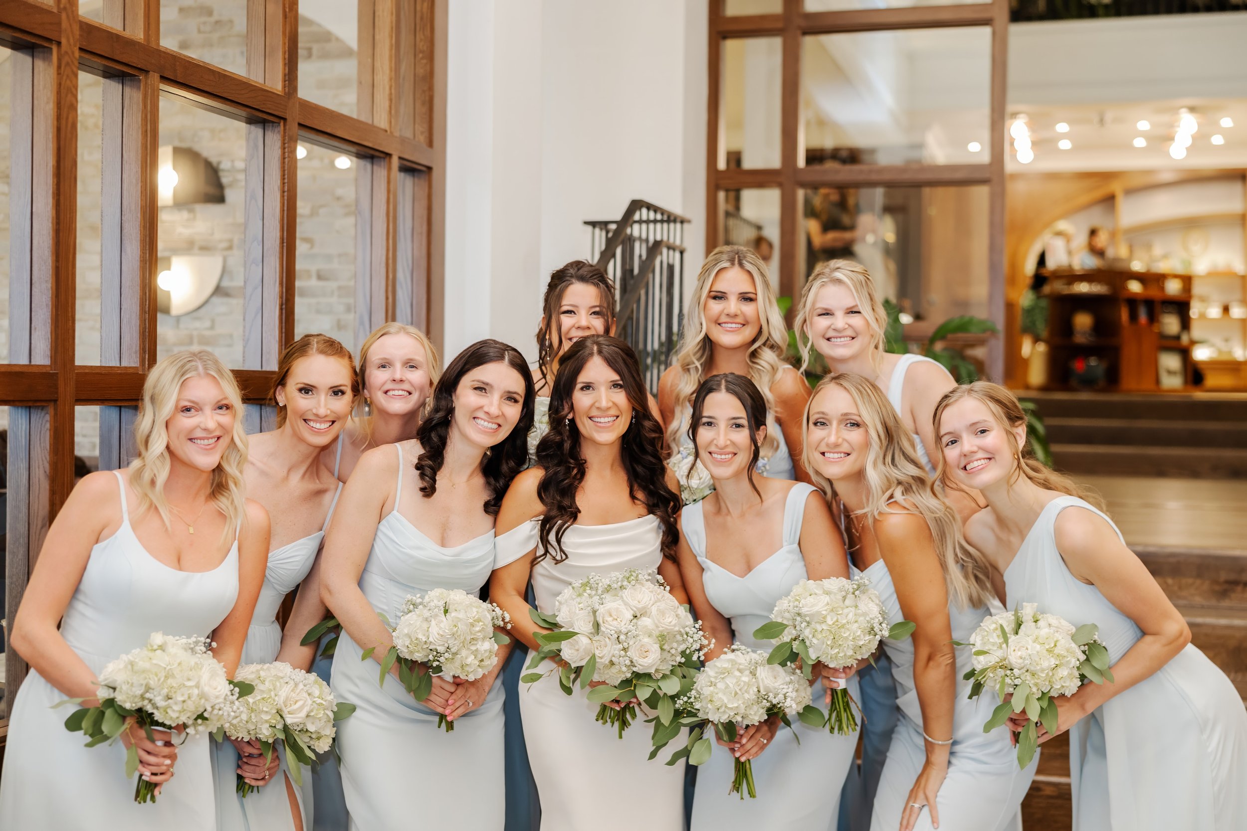 A group of eleven women dressed in matching light blue bridesmaid dresses, holding white bouquets, gathered indoors at a wedding venue with wooden decor and stairs in the background.