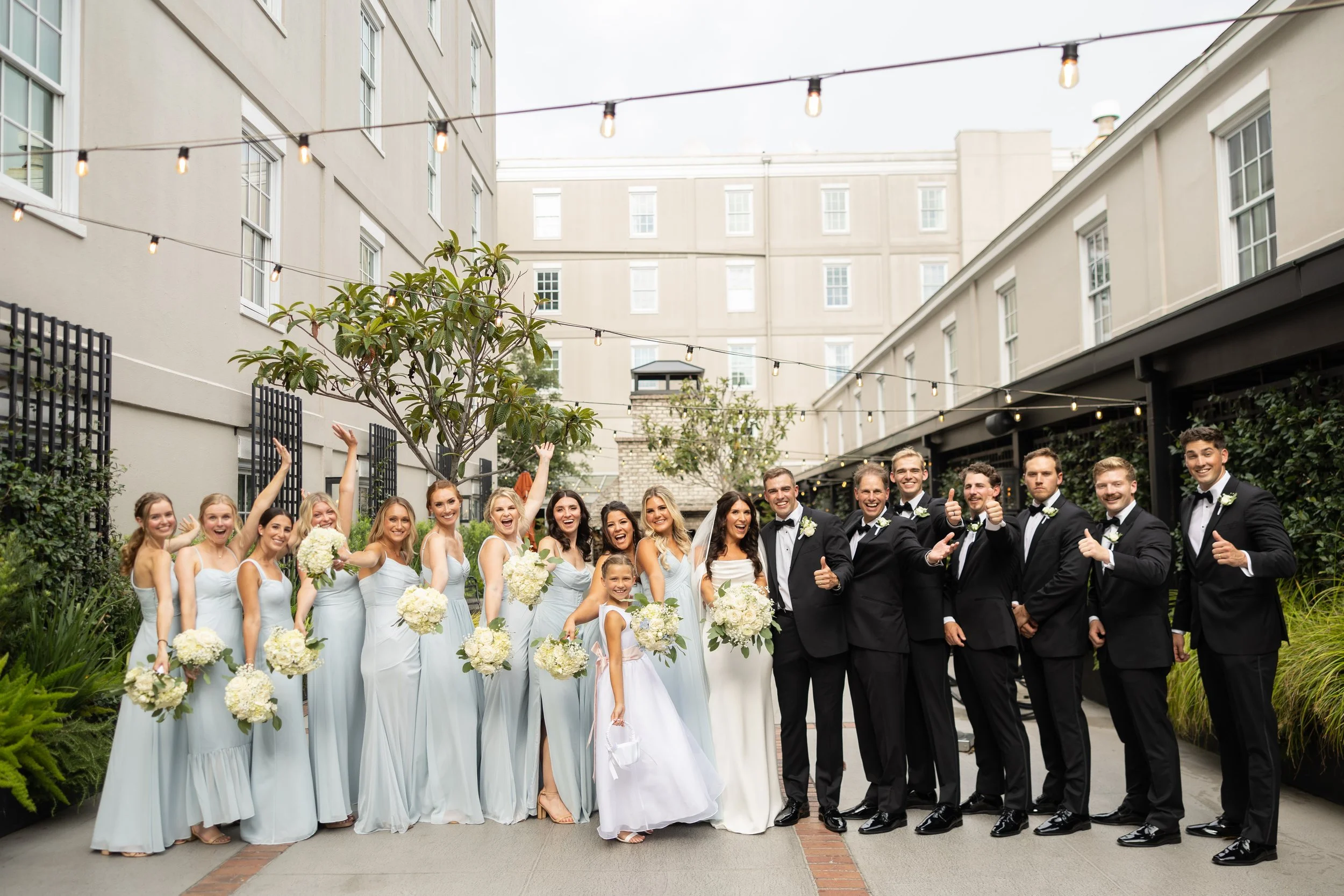 Group of wedding guests, including bridesmaids in light blue dresses and groomsmen in black tuxedos, posing outdoors with smiles, some giving thumbs up, under string lights in an urban courtyard.