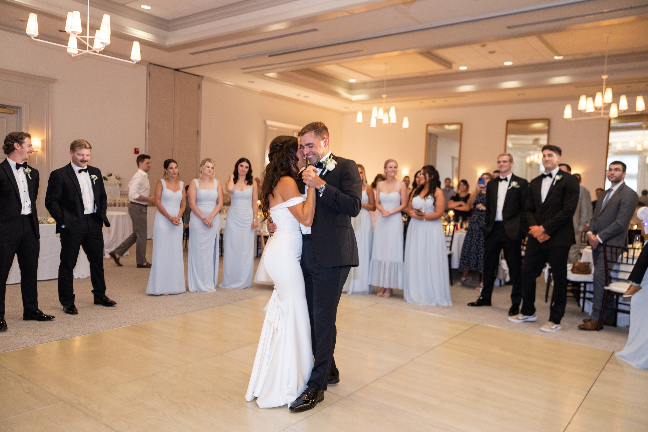 A bride and groom are dancing in the center of a wedding reception with guests watching around them. The bride wears a white off-the-shoulder gown, and the groom is in a black tuxedo. Guests are dressed in formal attire, some in black tuxedos and oth