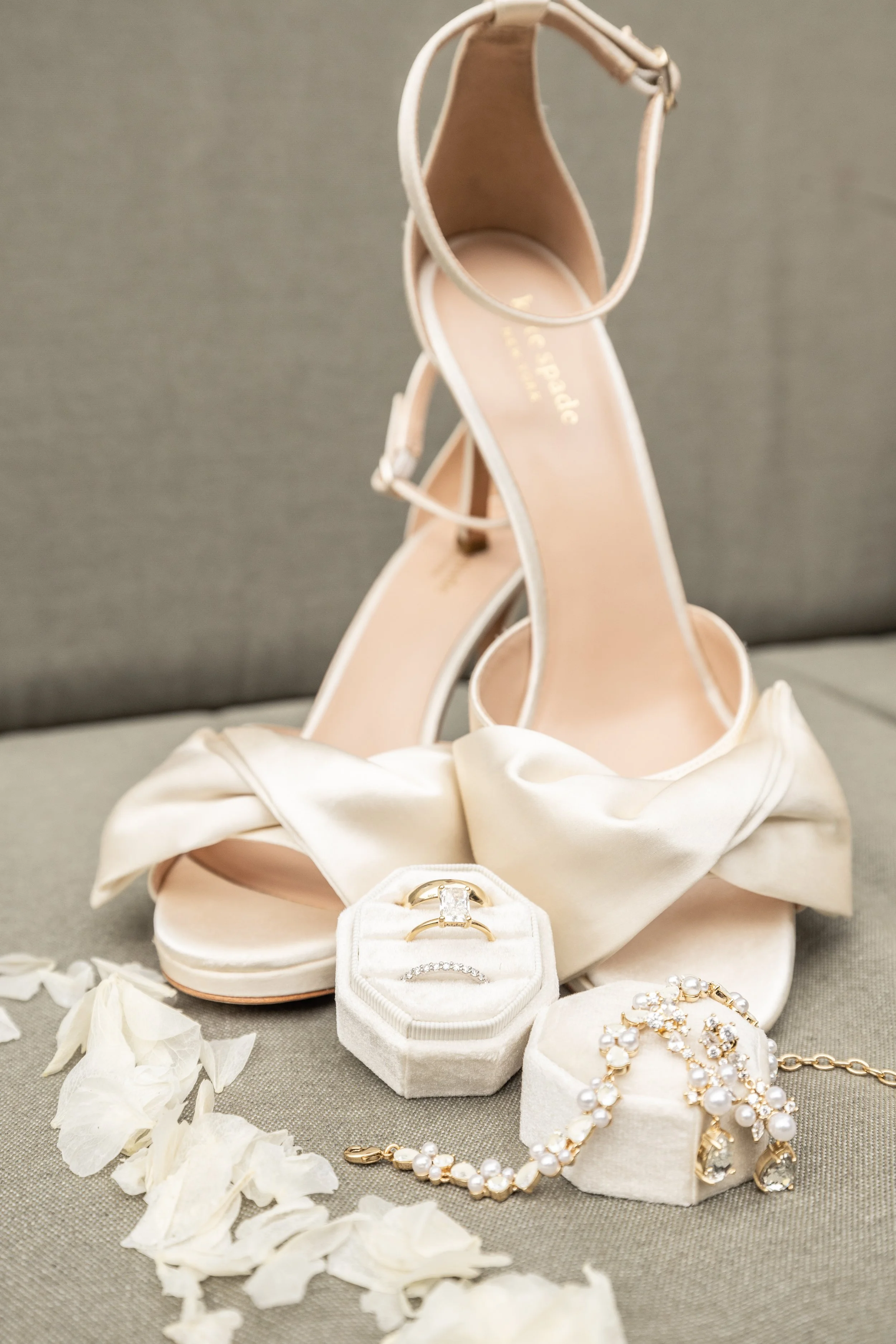 A pair of beige high-heeled sandals with ankle straps, surrounded by wedding jewelry including rings, a pearl bracelet, and a pearl necklace, along with white flower petals.