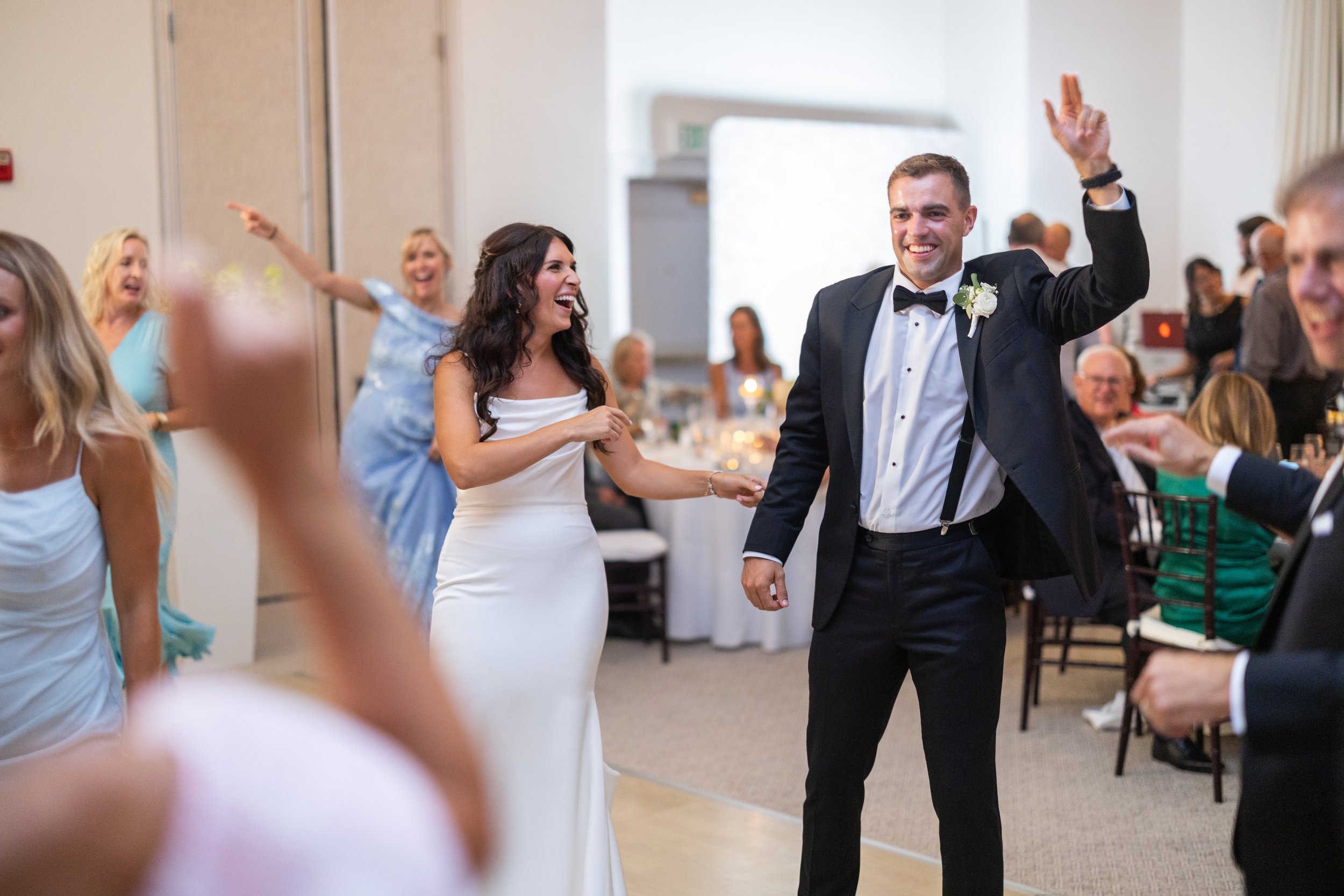 A joyful wedding reception scene with a bride in a white gown and a groom in a tuxedo dancing, surrounded by guests smiling and celebrating.